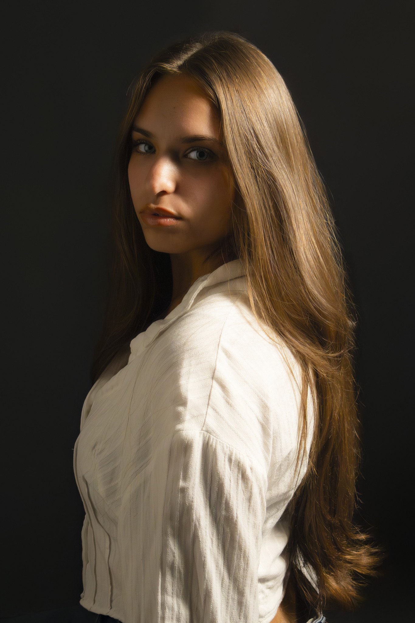 A young woman with long, flowing brown hair and blue eyes, wearing a white shirt, standing against a dark background.