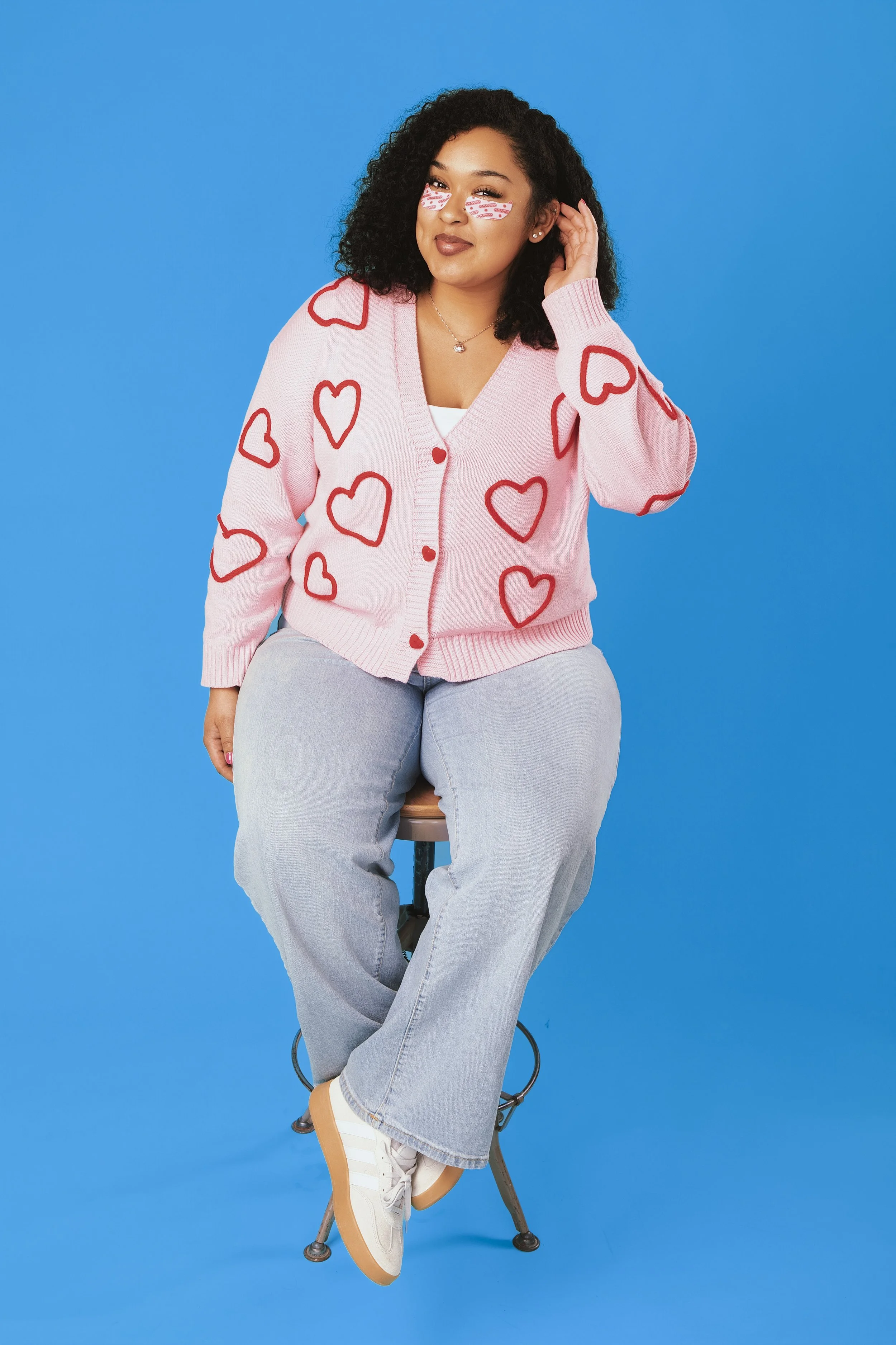 Young woman with curly black hair sitting on a stool against a blue background. She is wearing a pink cardigan with red hearts, gray jeans, and white sneakers. She has bandages with red lipstick print on her right eye, a necklace, earrings, and is sm
