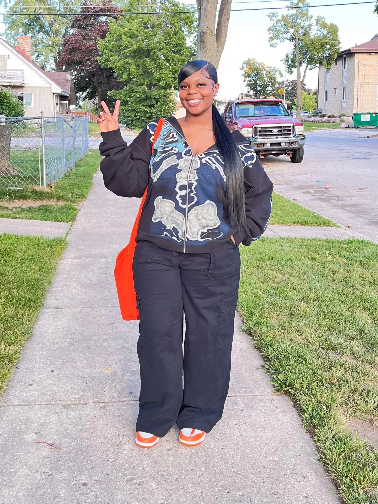 A young woman standing on a sidewalk in a neighborhood, making a peace sign with her right hand. She's smiling and wearing a black and gray hoodie with a skull design, black pants, and orange and white sneakers. A red bag hangs from her shoulder. Behind her, there are trees, houses, a red pickup truck, and a green trash bin.