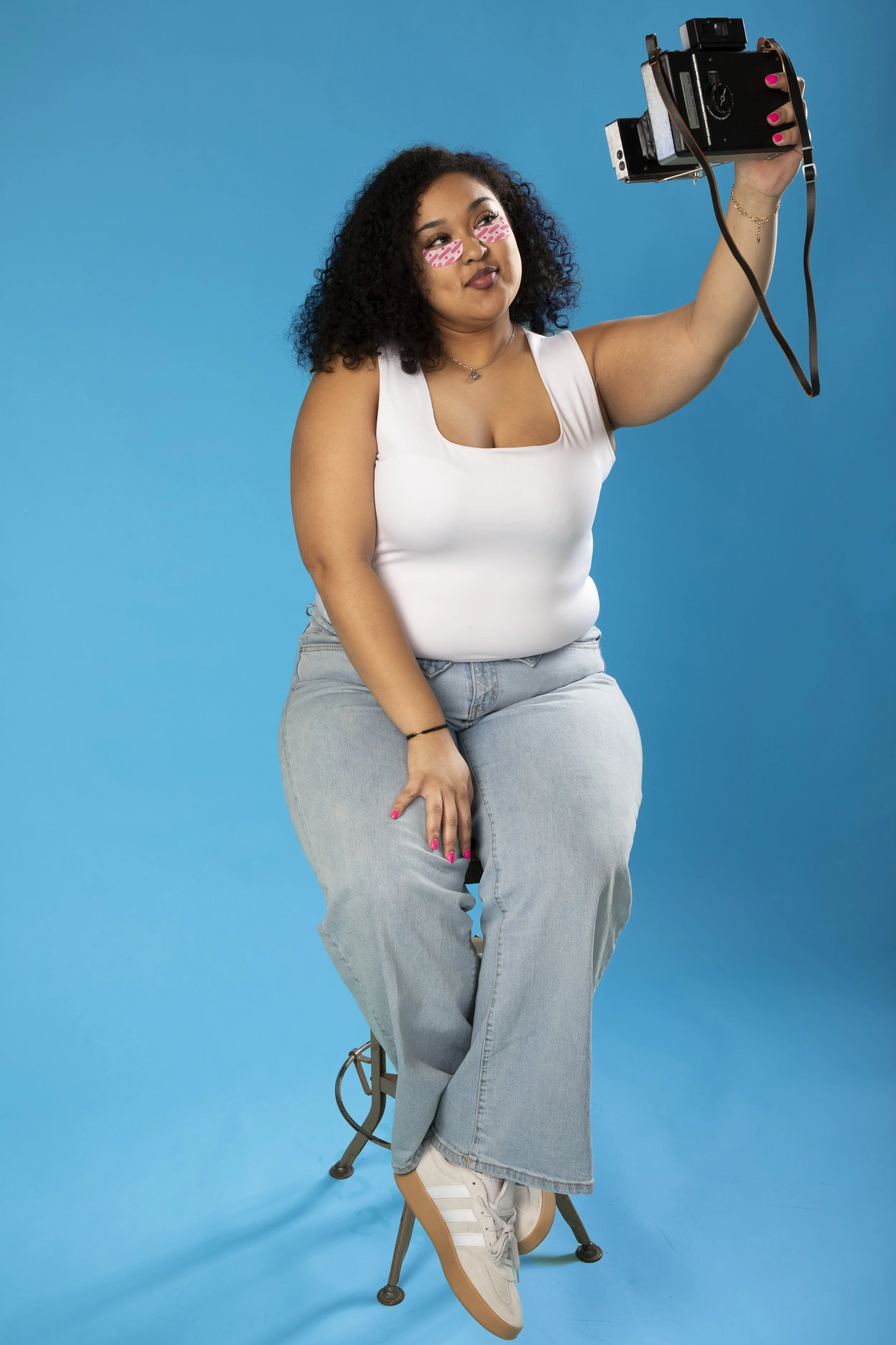 Young woman with curly hair, wearing pink eye masks, white tank top, and light blue jeans taking a selfie with a vintage camera against a blue background.
