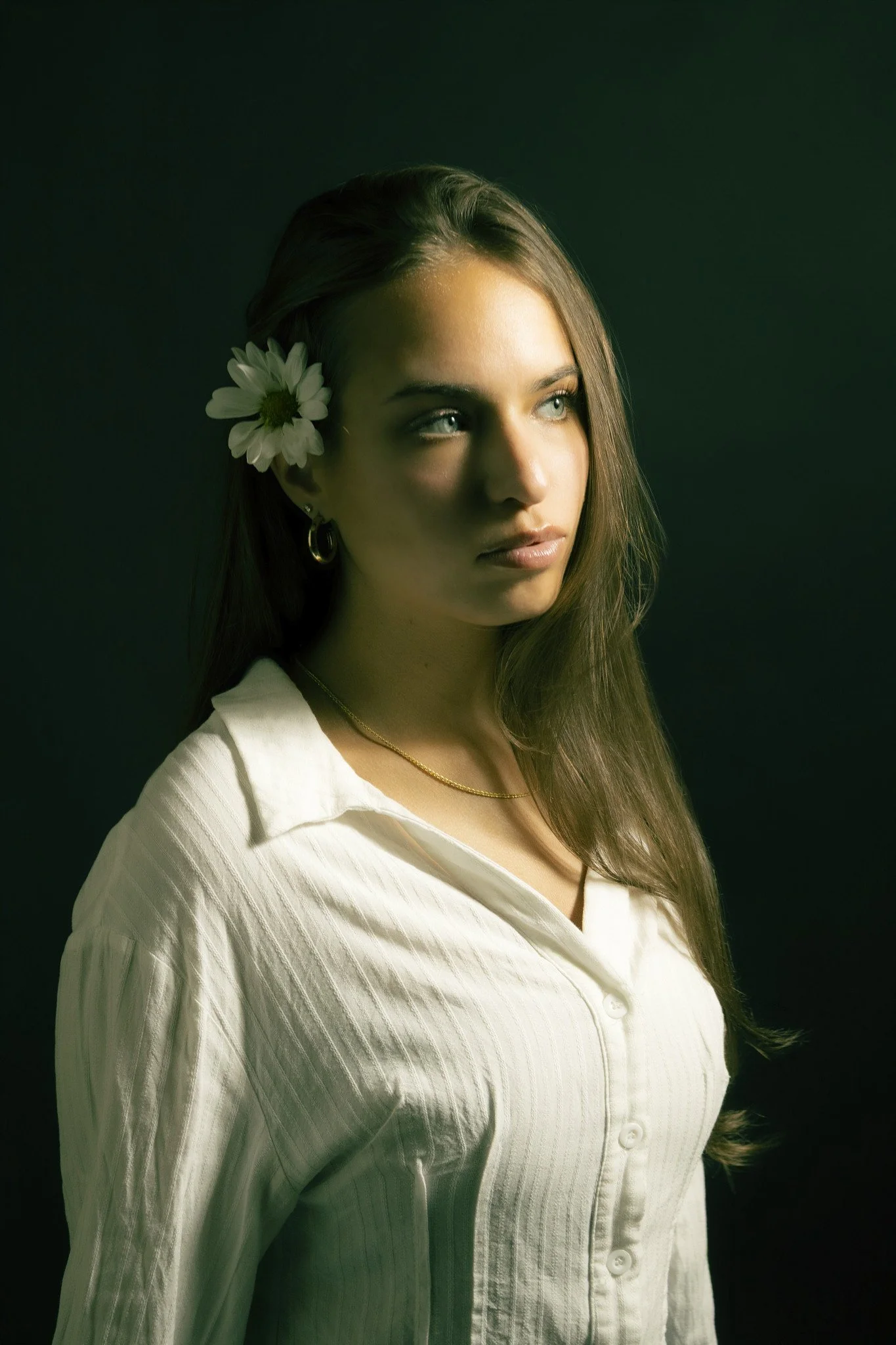 A young woman with long brown hair, light skin, and blue eyes wearing a white button-up shirt, a gold necklace, hoop earrings, and a daisy flower in her hair, looking to her right against a dark background.
