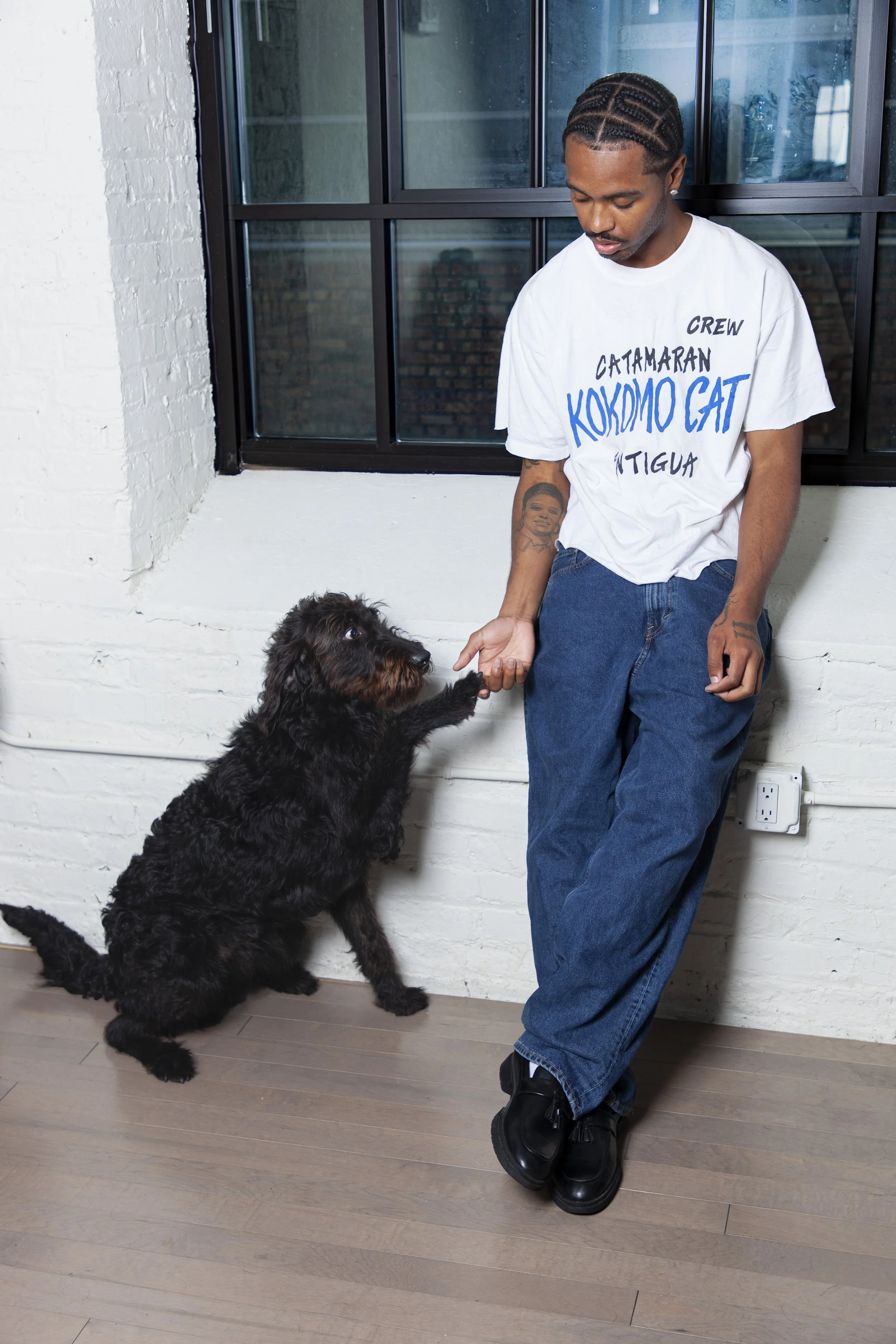 A man with braided hair, wearing a white T-shirt with blue and black writing, jeans, and black shoes, is standing indoors near a large window with a brick wall visible outside. He is interacting with a large black dog, possibly a Bernese Mountain Dog