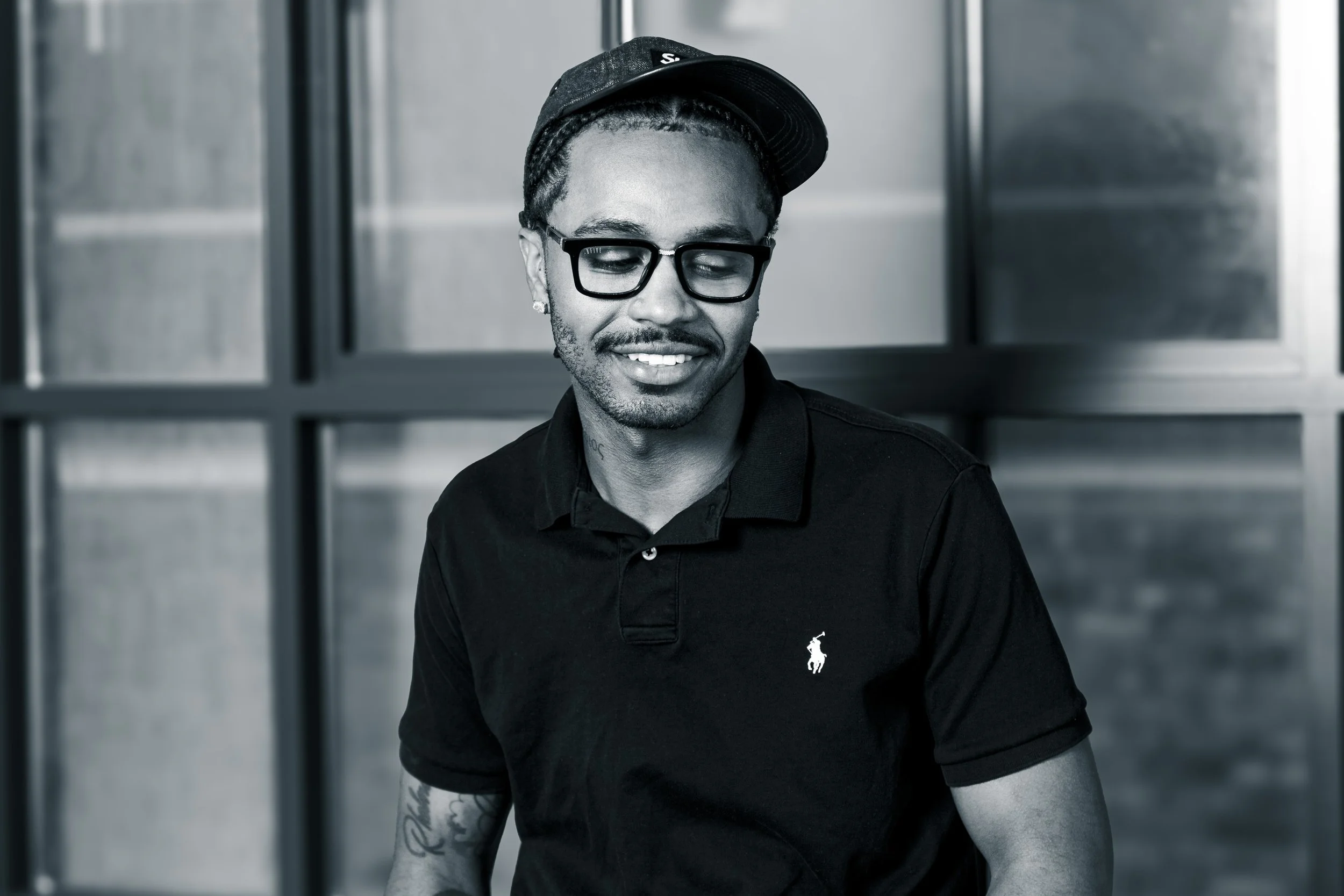 A young man with braided hair, glasses, a baseball cap tilted sideways, and a tattoo on his arm, smiling and looking down, wearing a black polo shirt with a small logo, standing in front of a textured wall.