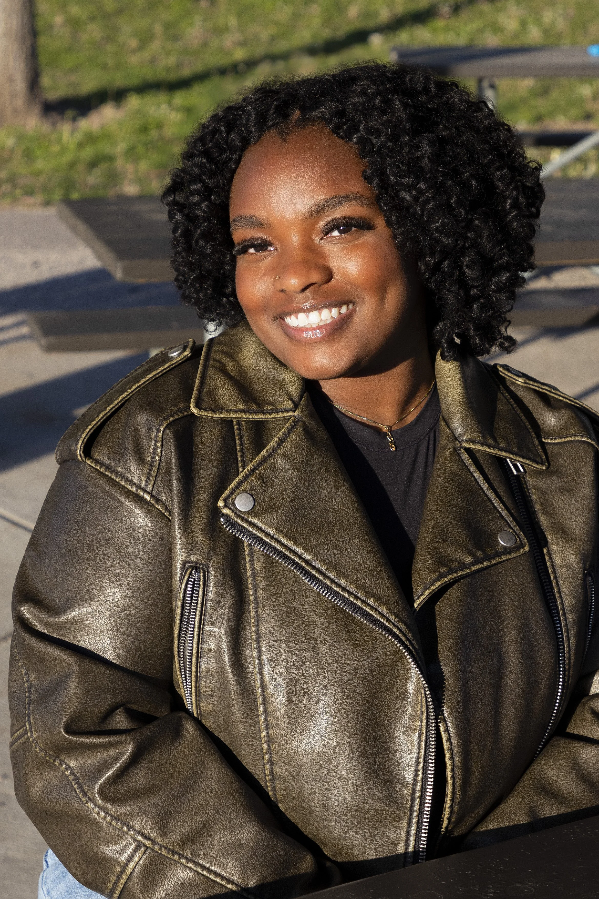 A woman with curly black hair smiling outdoors in natural light, wearing a brown leather jacket and a small gold necklace.