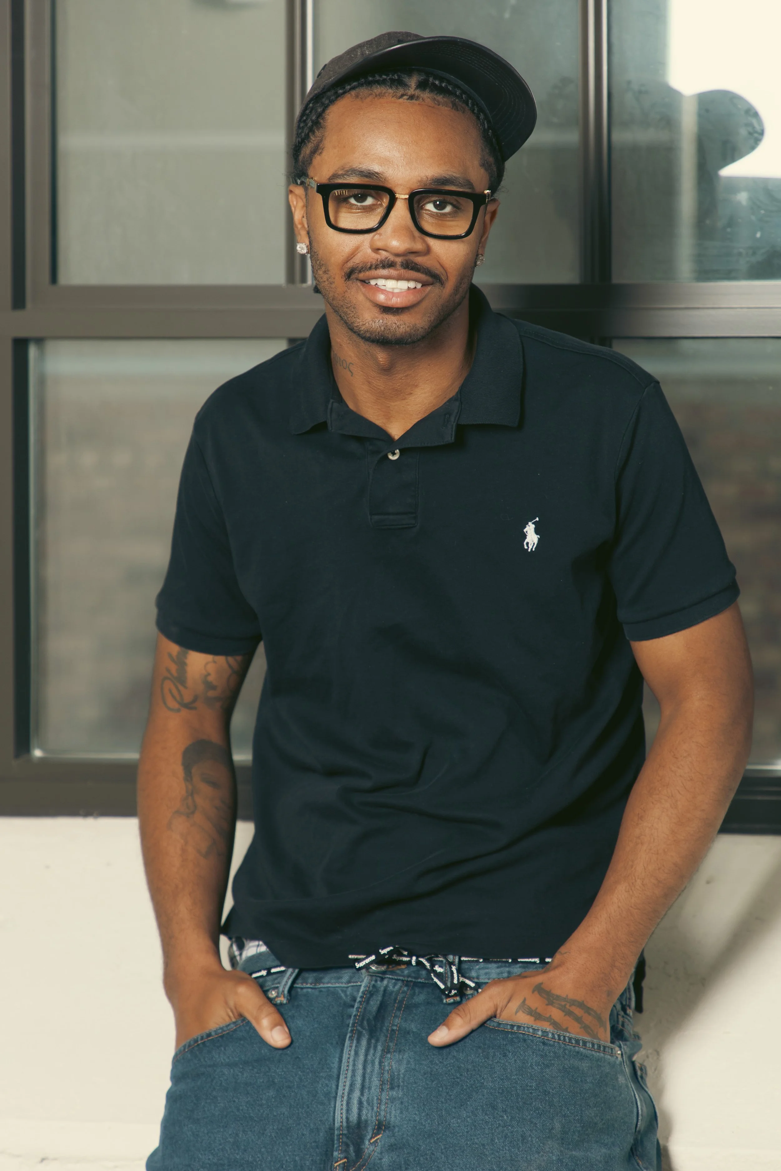Young man with glasses, earrings, a black cap, and a black polo shirt, smiling and leaning against a glass wall.
