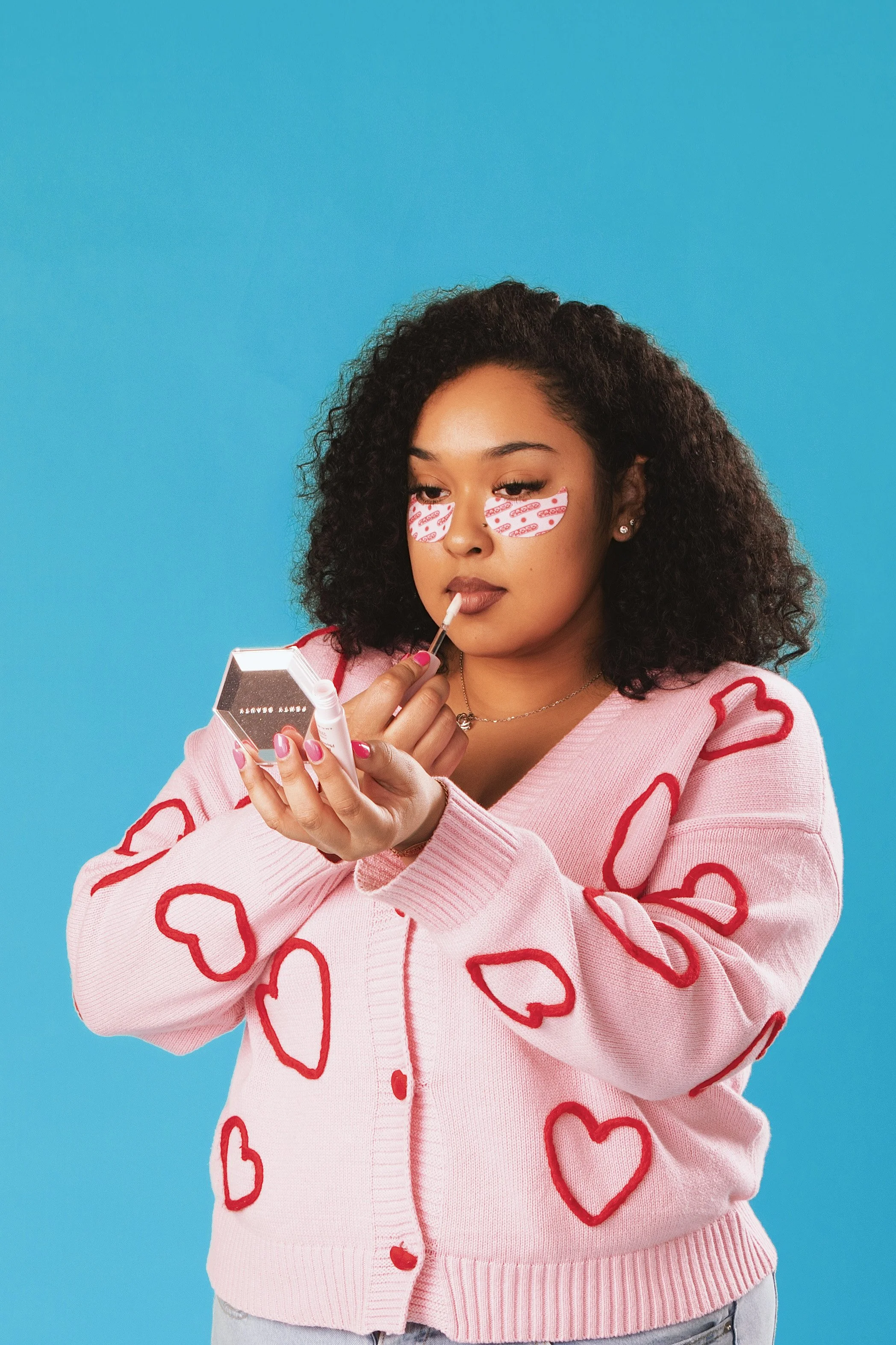 A woman with curly hair applying lip gloss in front of a turquoise background, wearing a pink sweater with red heart outlines.