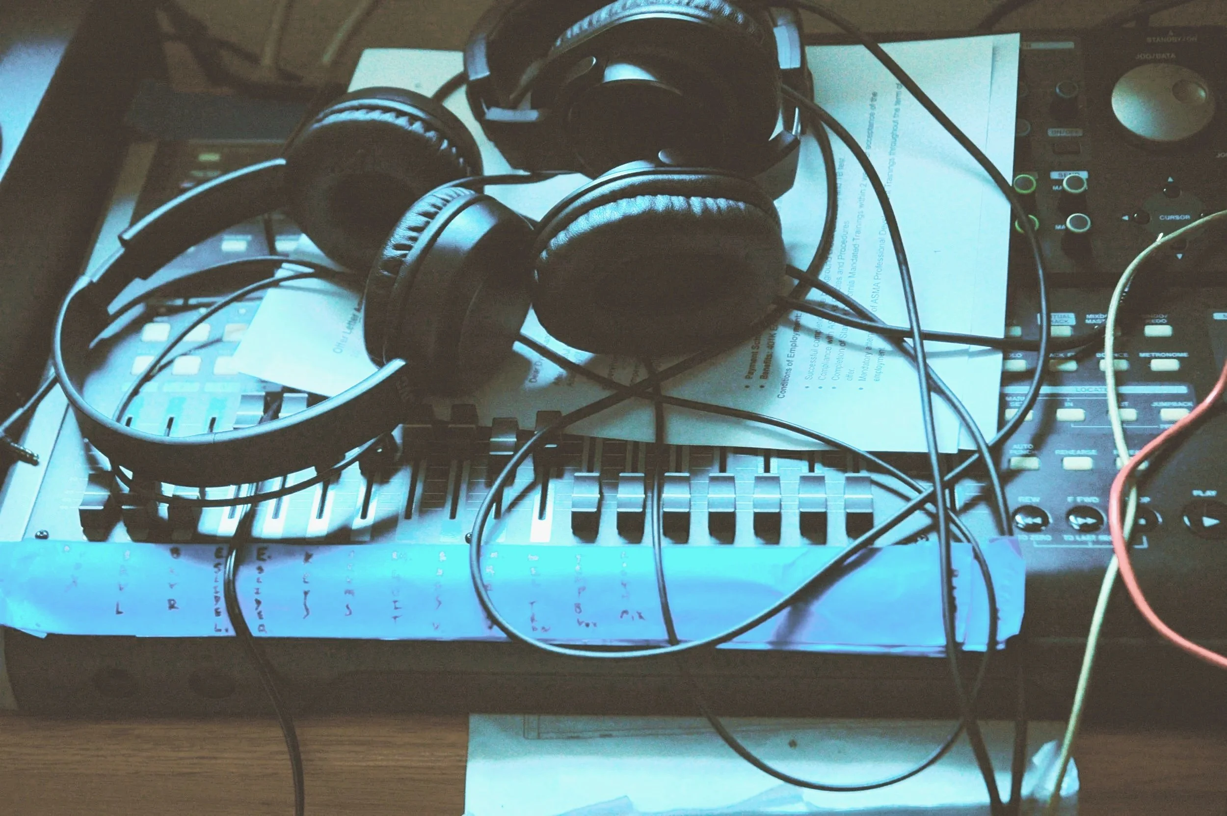 A pair of black headphones resting on a sheet of paper with a printed document, surrounded by audio equipment and tangled wires on a desk.