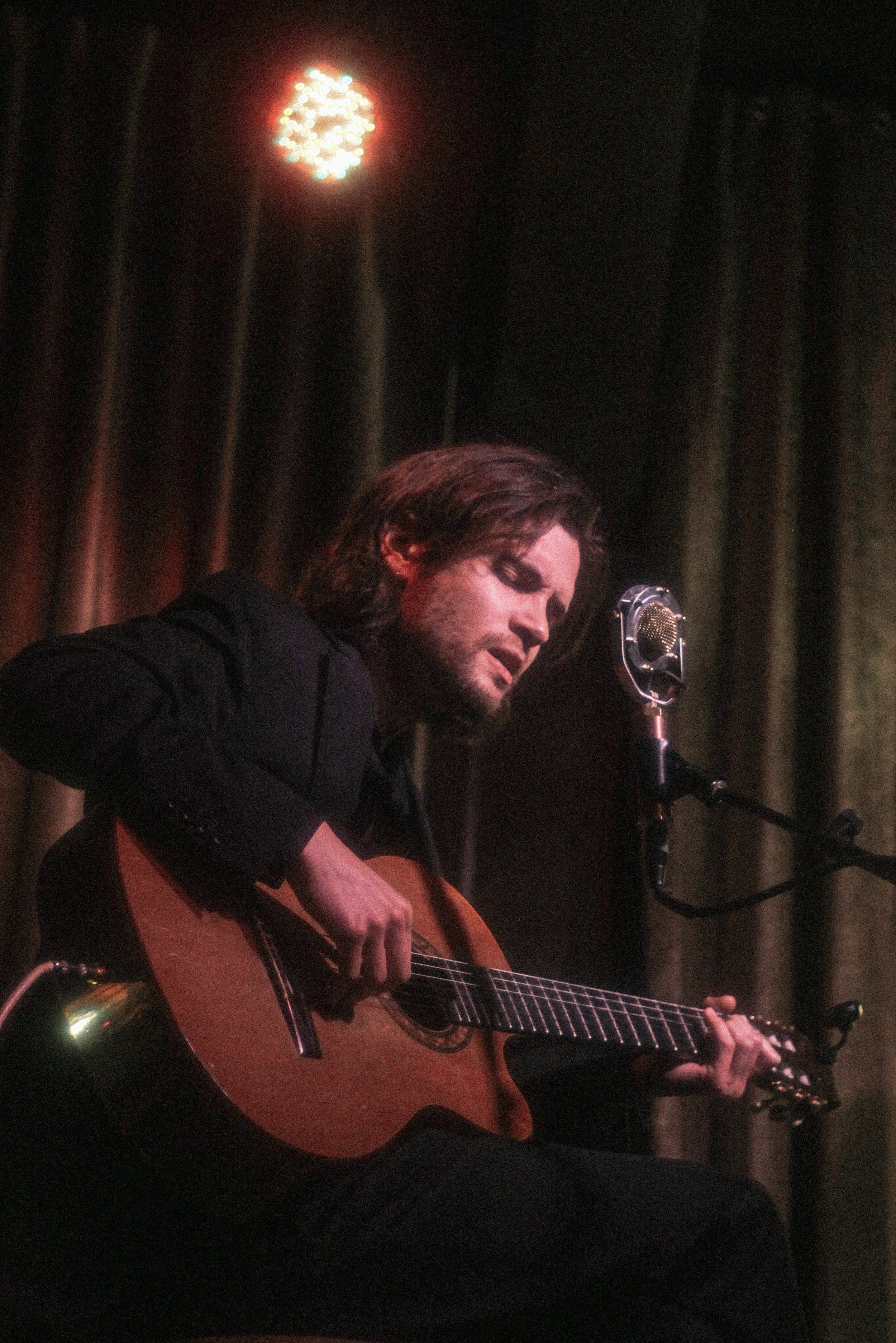 A man with longer brown hair playing an acoustic guitar on stage in front of a dark curtain with red lighting.