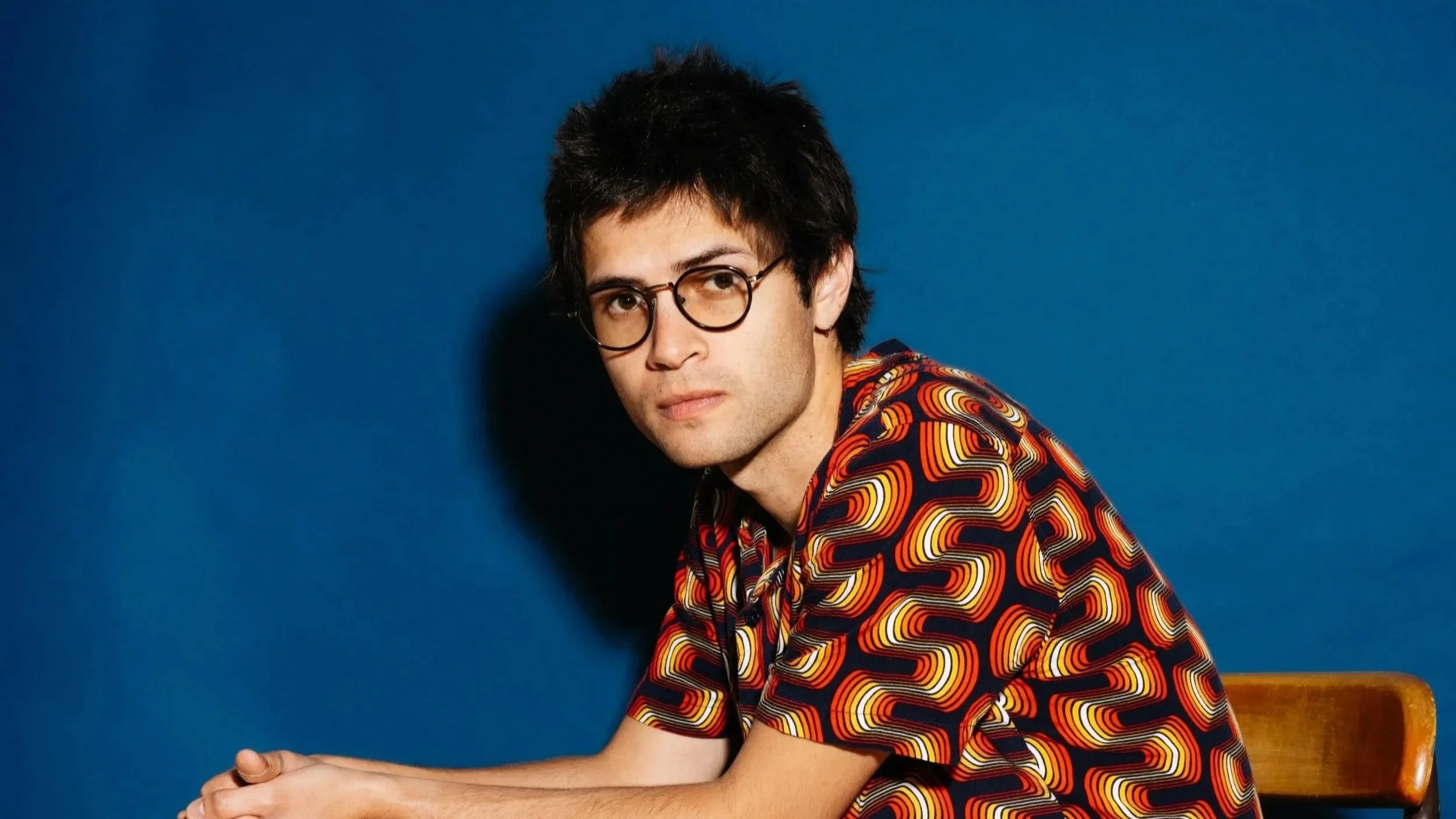 A young man with dark, curly hair and glasses posed against a solid blue background. He is wearing a multicolored, patterned shirt and is seated on a wooden chair.