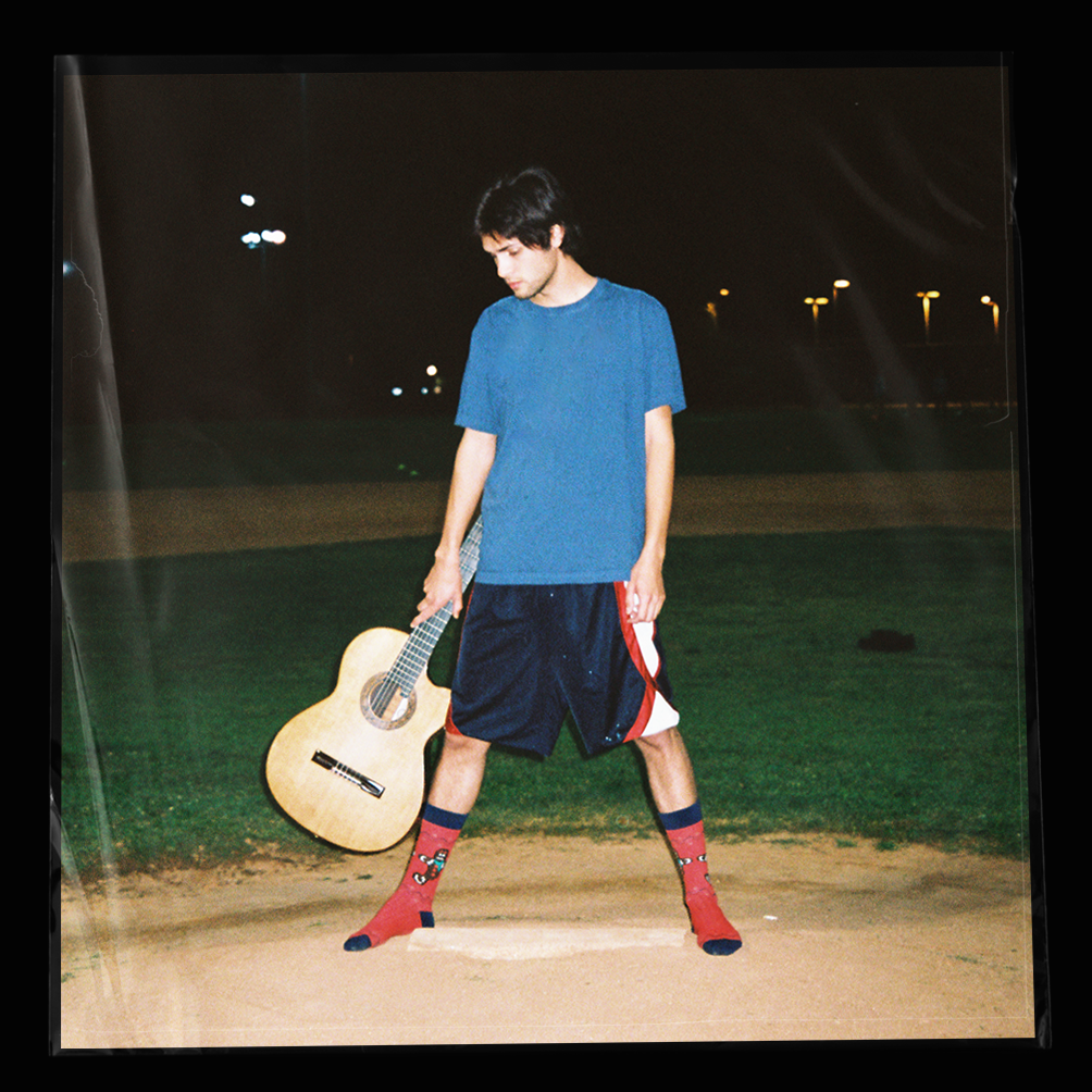 Young man standing on a baseball field at night, holding an acoustic guitar, wearing a blue t-shirt, shorts, and colorful socks.