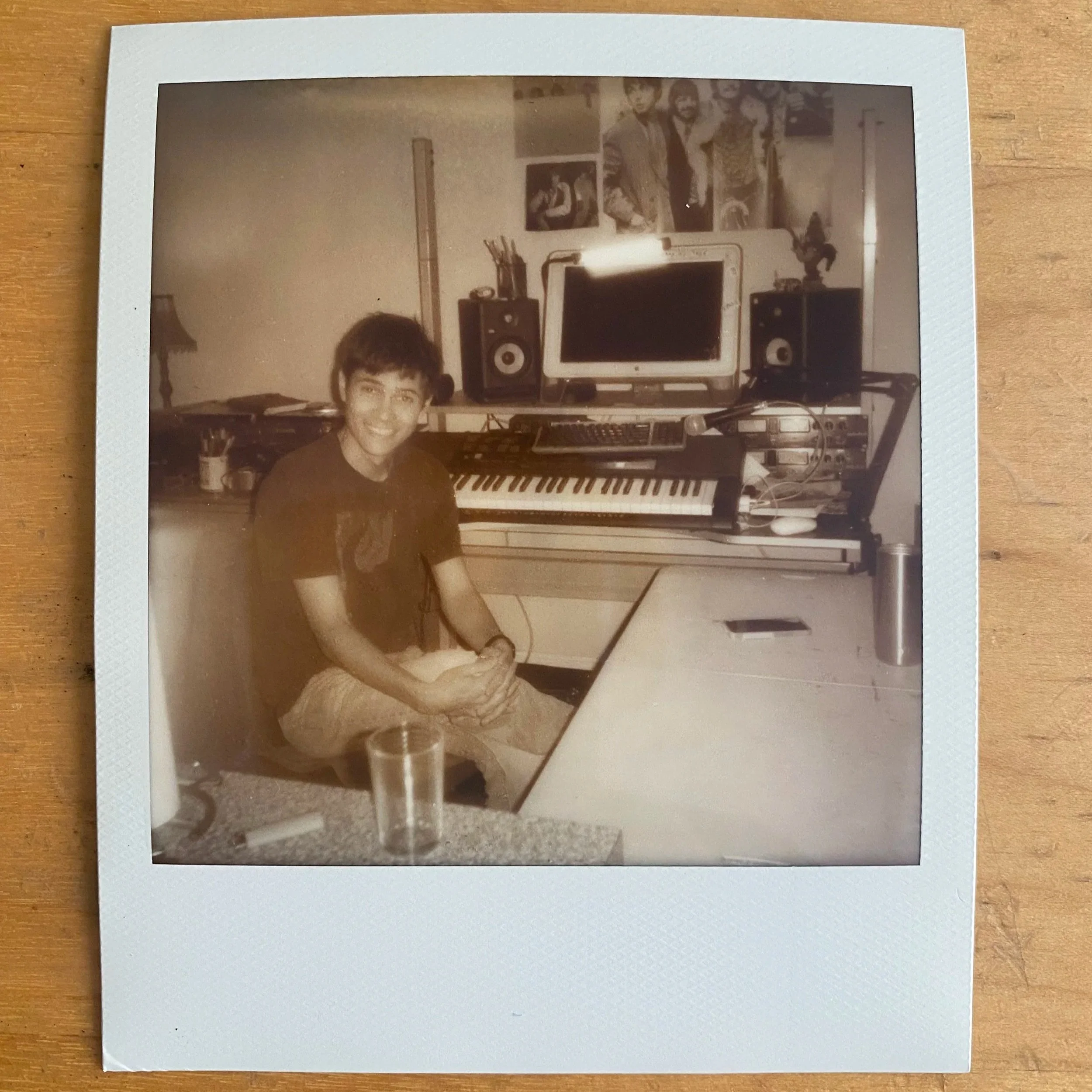 A young person with short hair smiling, sitting at a music production setup in a room decorated with framed pictures and posters on the wall.