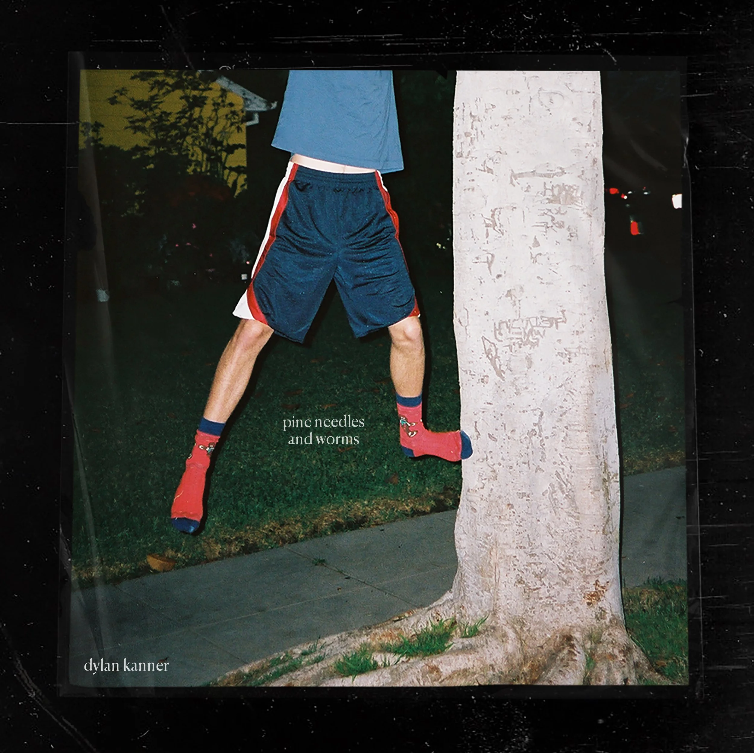 A man wearing a blue jacket, navy shorts with red and white stripes, red socks, and black shoes stands outdoors on grass with one hand on his head, next to a white textured wall at night. In the background, trees and a faintly lit house are visible, with the text "dylan kanner" at the bottom left of the image.