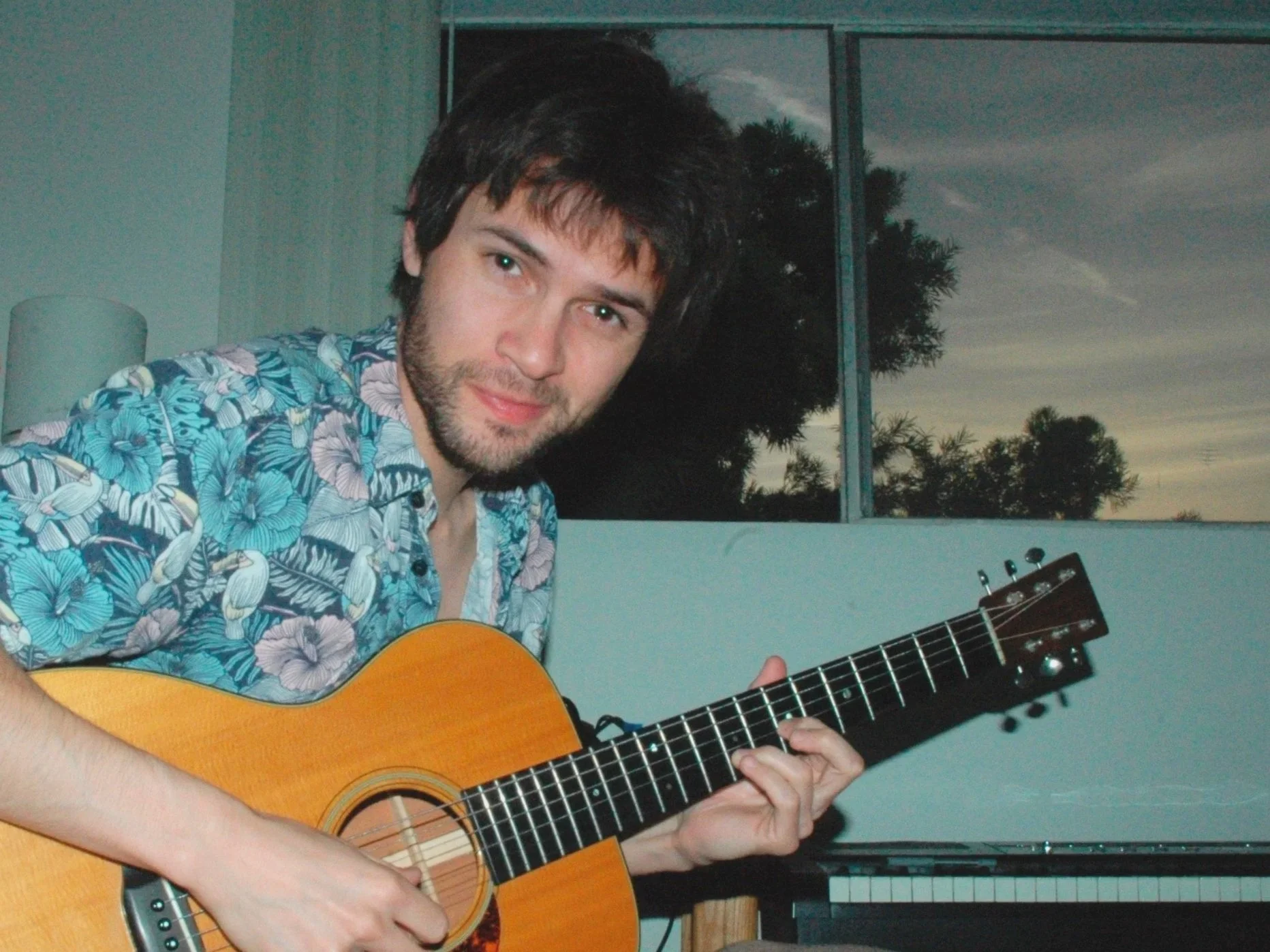 A young man with dark hair and a beard playing an acoustic guitar indoors, with a background of large windows showing trees and a dusk sky.