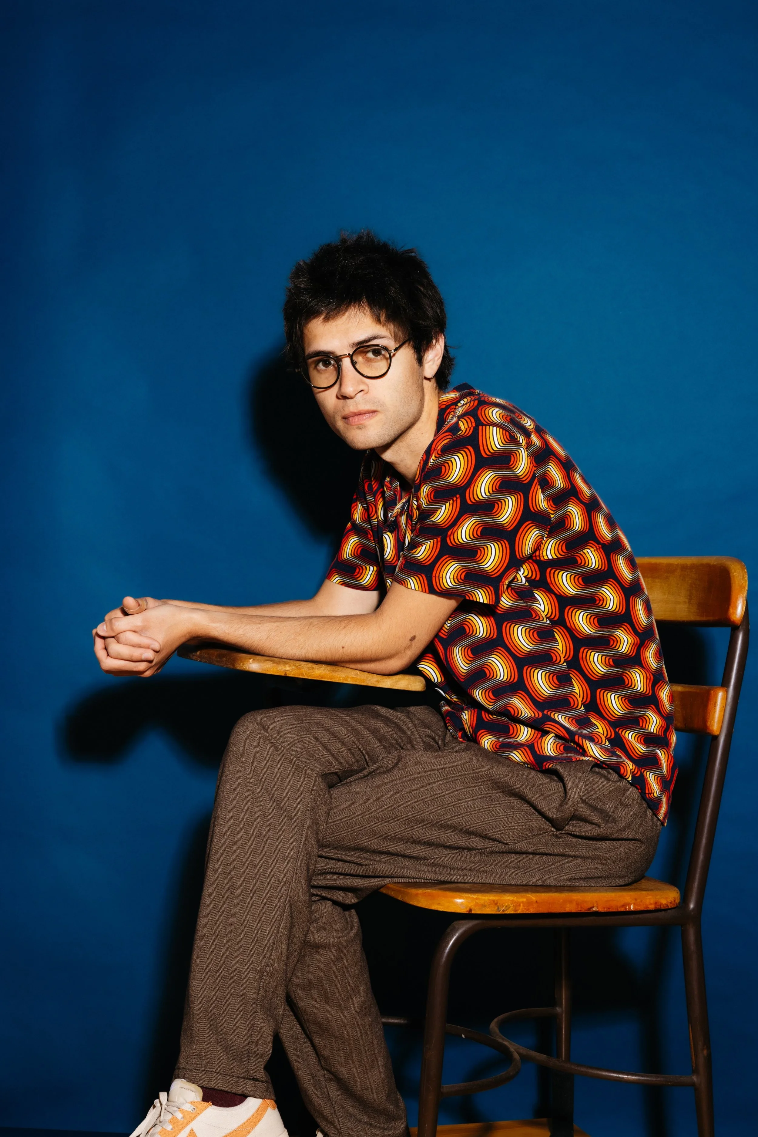 A young man with black hair and glasses sitting on a wooden chair against a blue background wearing a colorful, patterned short-sleeve shirt, brown pants, and white sneakers with orange and yellow accents.