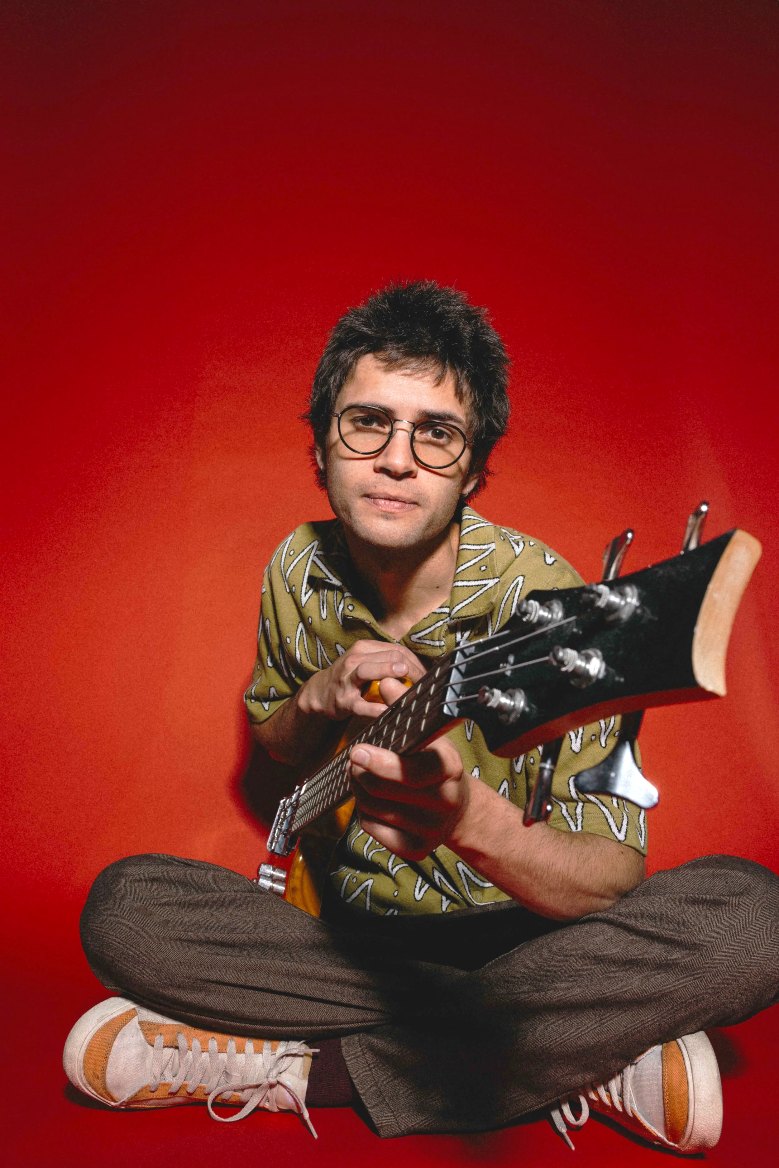 A young man with dark messy hair, glasses, wearing a patterned shirt, sitting cross-legged against a red background, holding an electric bass.