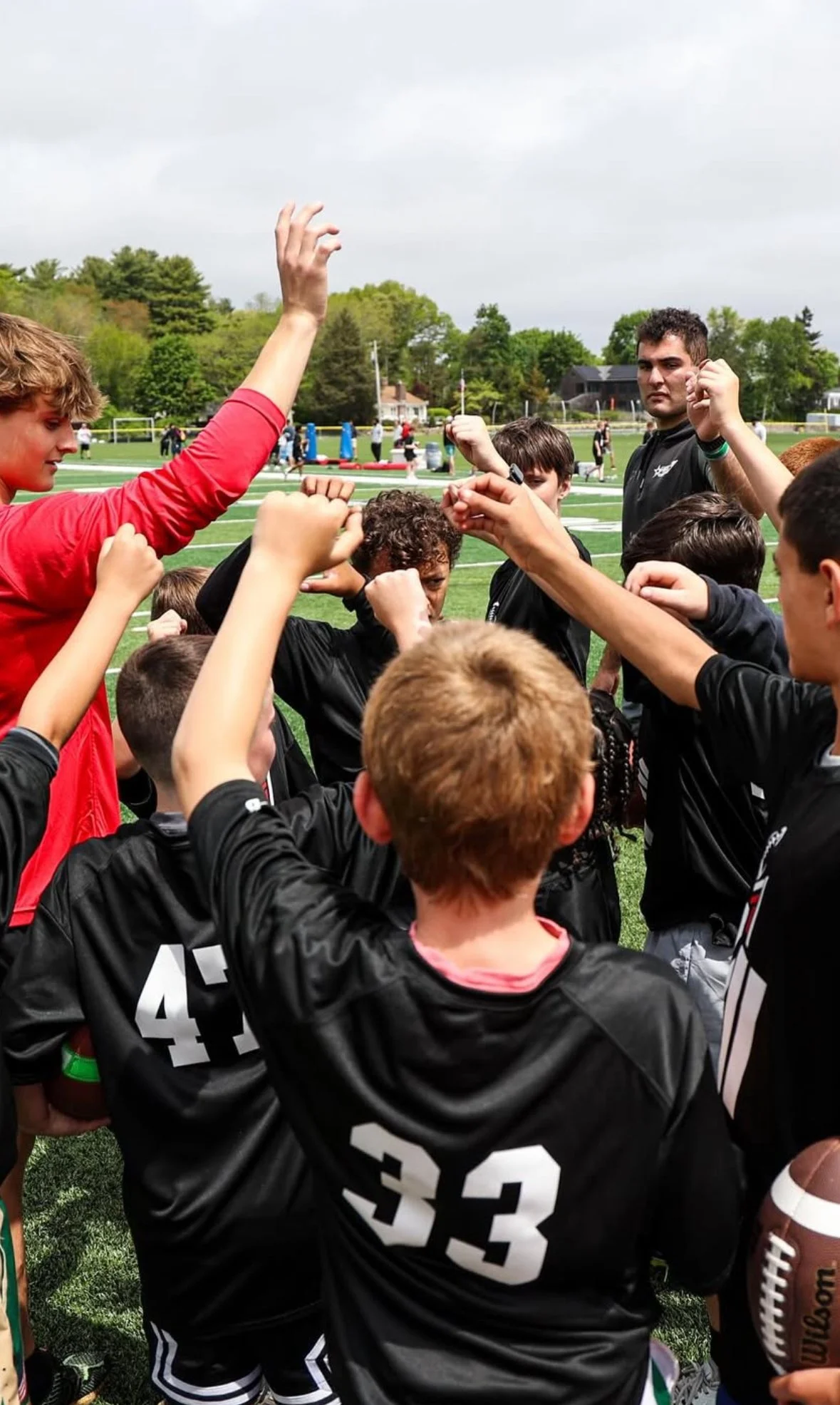 A group of young football players in black jerseys with white numbers, gathered in a huddle on a football field during daytime, with a coach or team leader giving instructions.