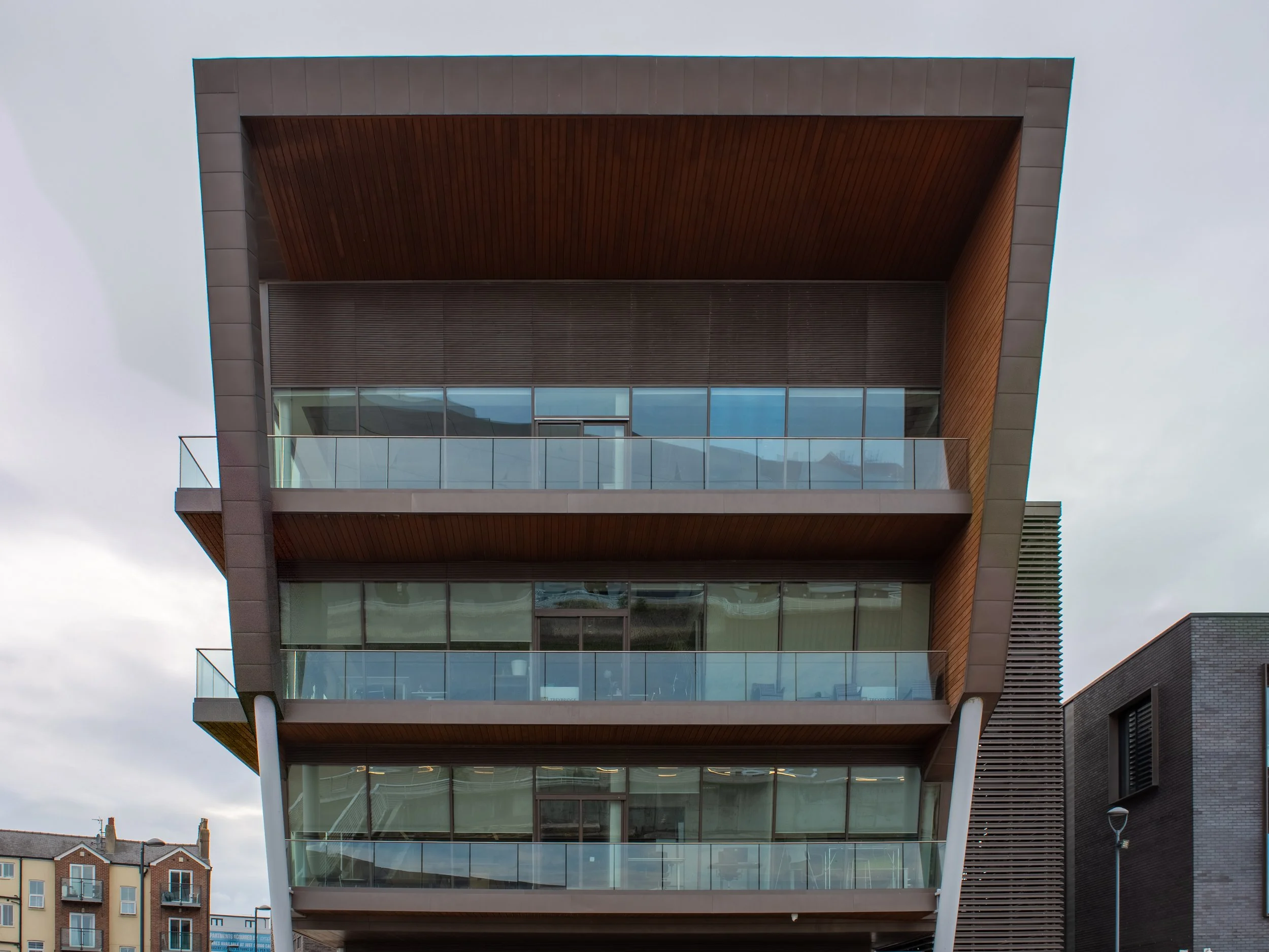 Modern multi-story building with extensive glass windows and wooden panel accents, set against a gray sky.