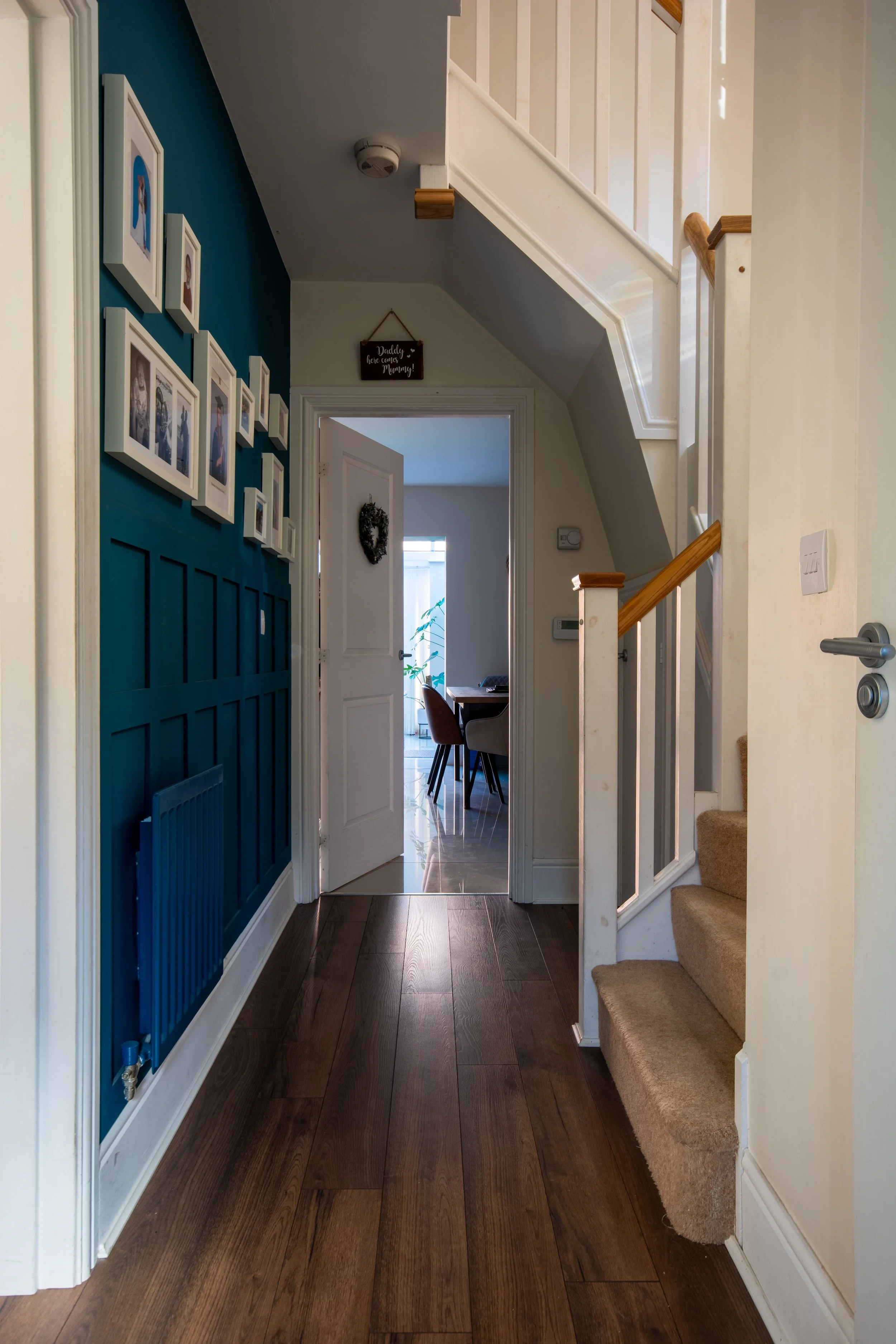Interior view of a hallway with wooden flooring, a staircase with carpeted steps on the right, a blue feature wall with framed pictures on the left, and an open door leading to a dining area with chairs and a window in the background.