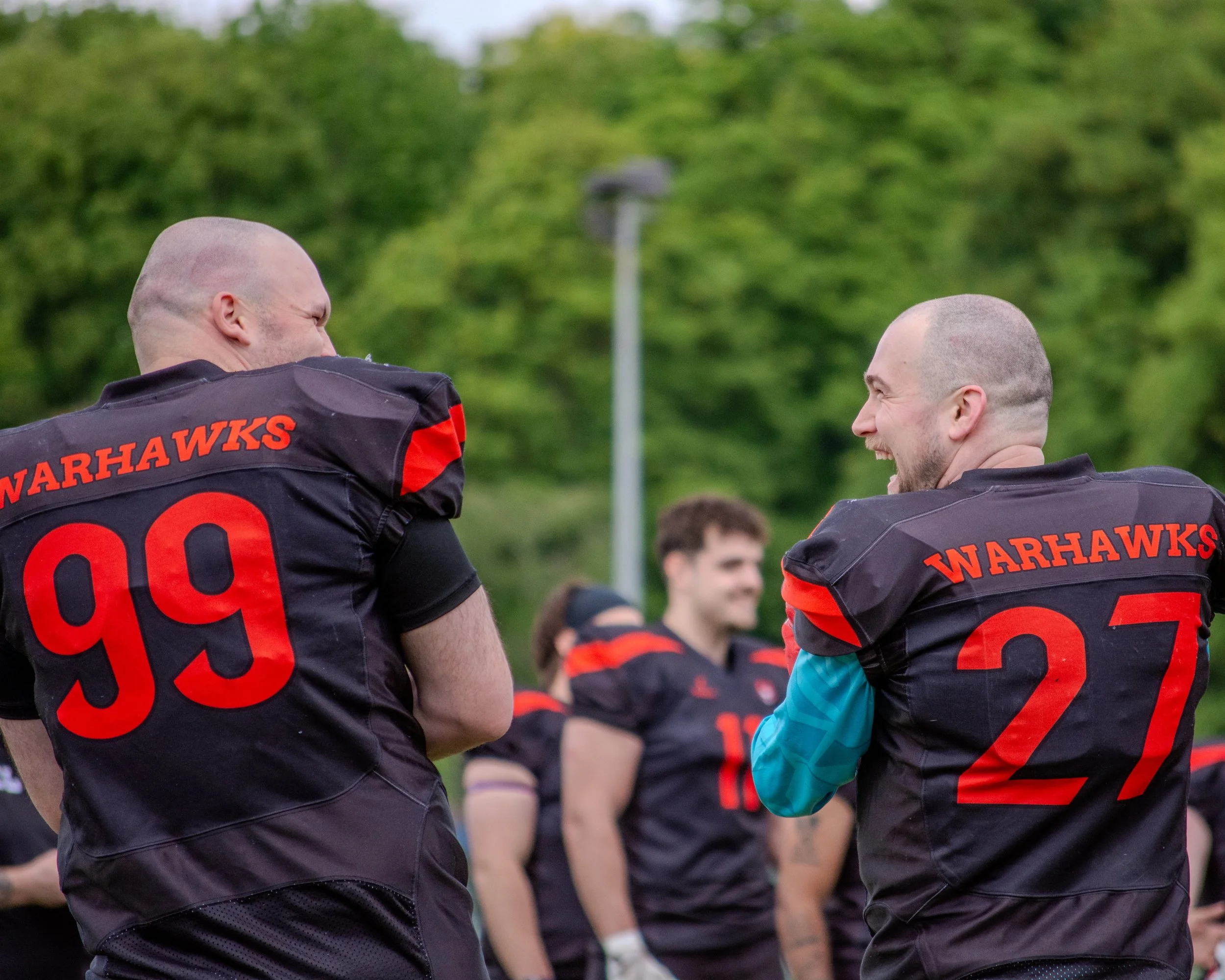 Two football players from the Warhawks team on the field, interacting with each other, wearing black and red uniforms with numbers 99 and 27, with green trees and blurred crowd in the background.