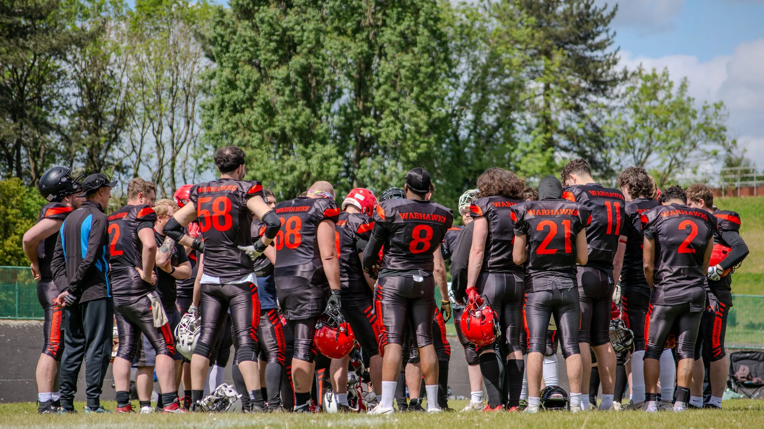 A group of football players and coaches gathered on a field, dressed in black and red uniforms, listening to a coach. Trees and blue sky are visible in the background.