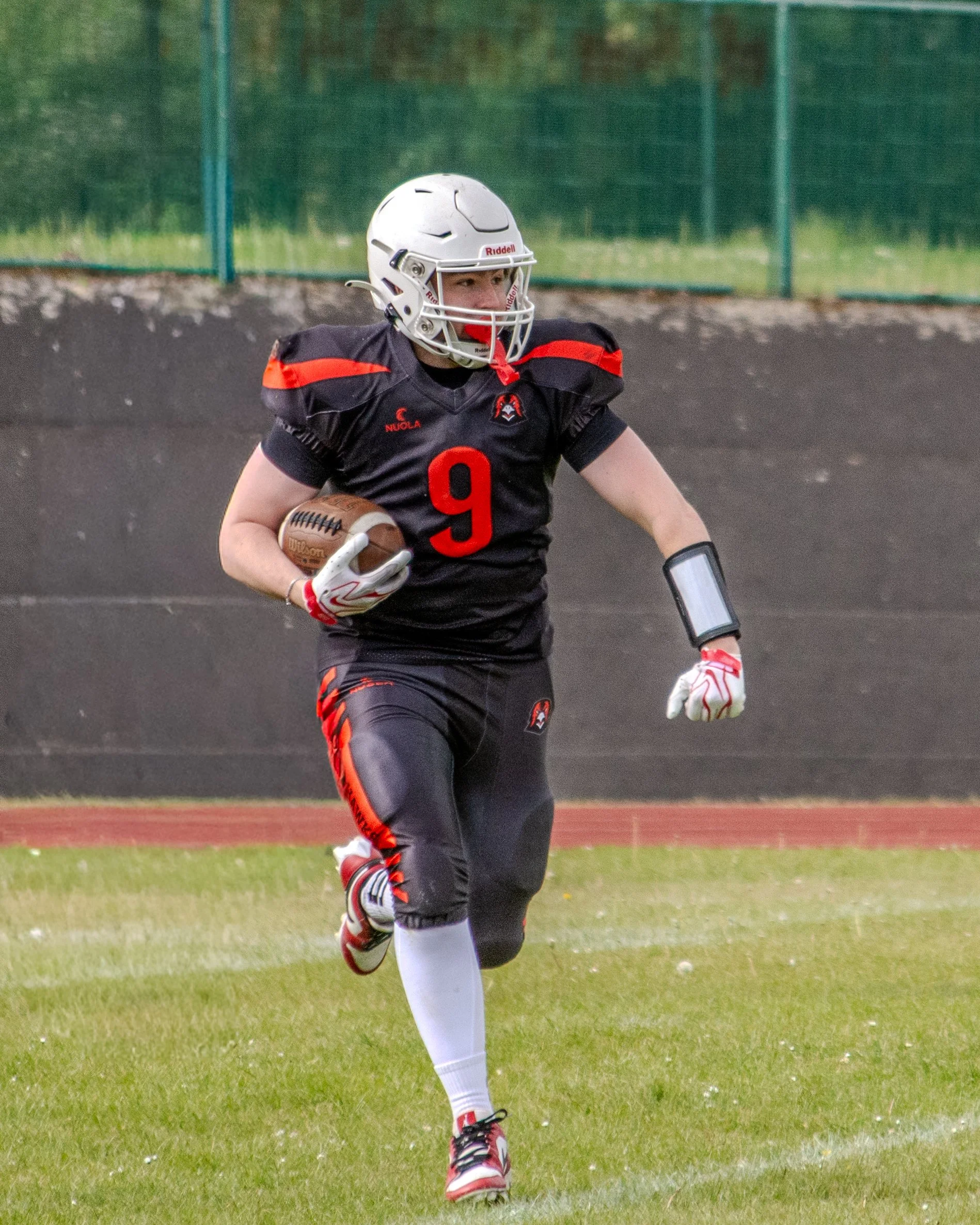 An American football player in a black and red uniform with the number 9, wearing a white helmet, red and white gloves, and running on a grassy field while holding a football.