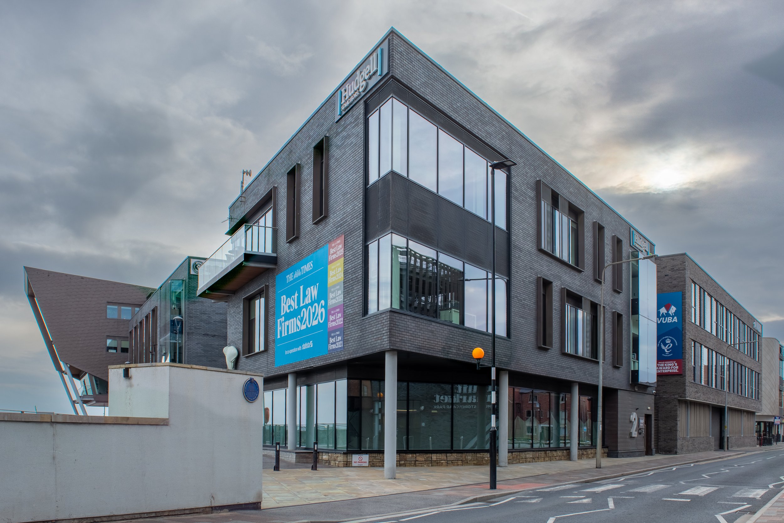 Modern multi-story office building with large glass windows and blue signage, situated on a city street under a cloudy sky.