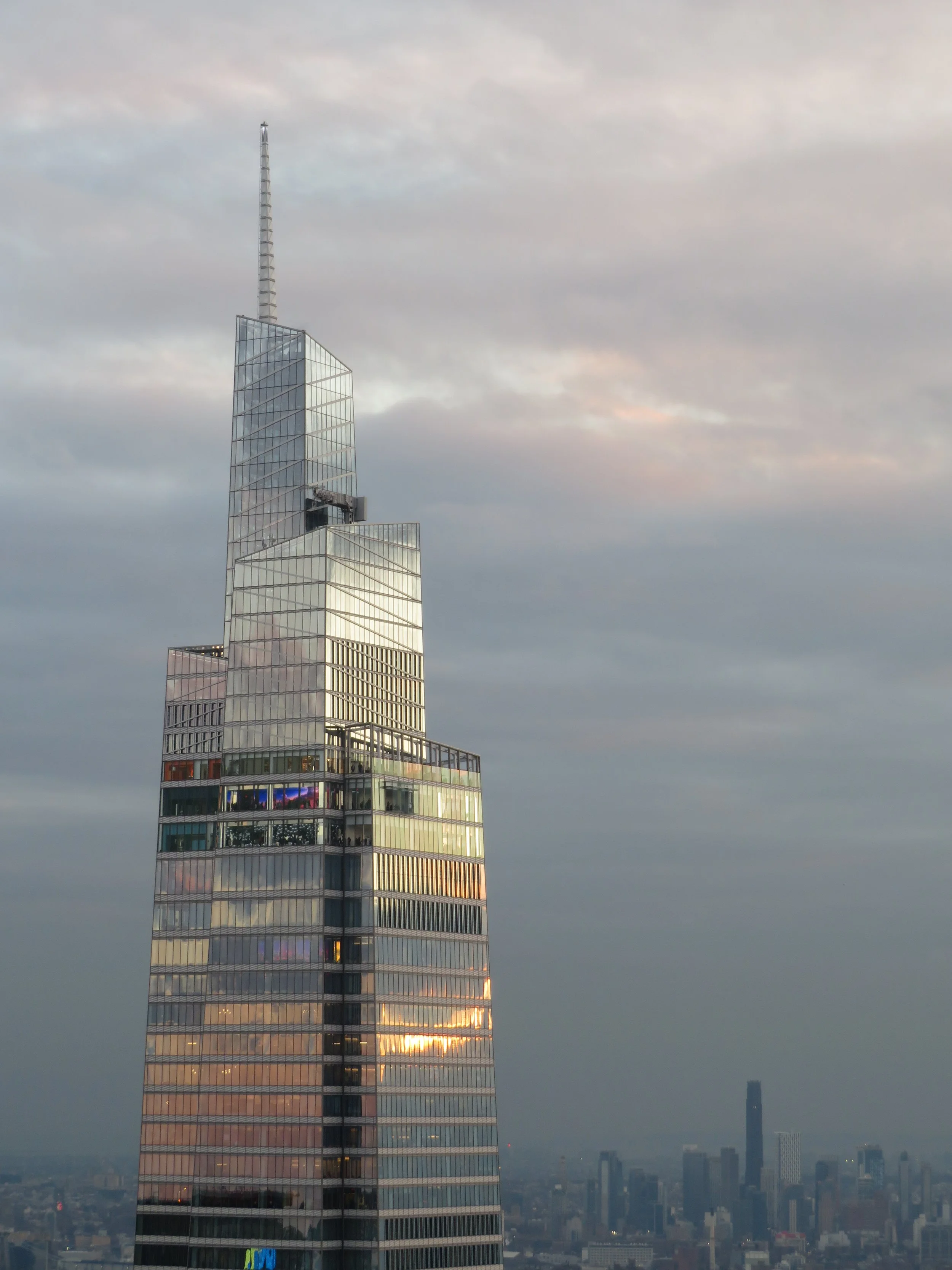 A tall modern skyscraper with a glass facade reflects the sky, with a city skyline and a few other tall buildings in the background, under a cloudy sky during sunset.