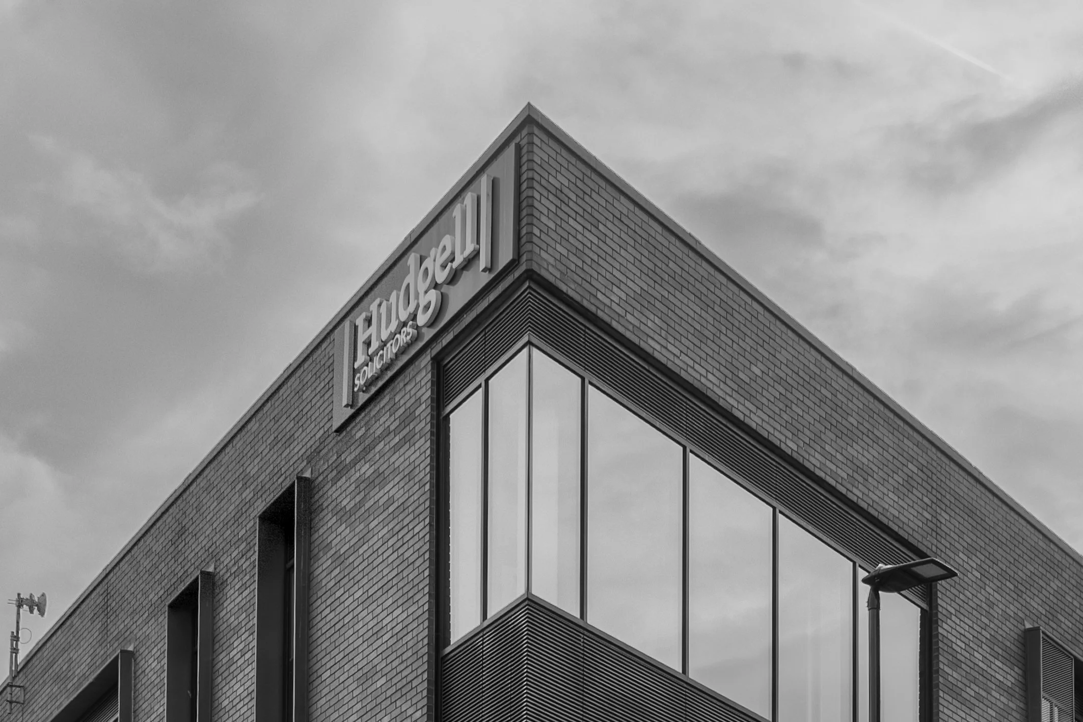 Black and white photo of a modern building with a sharp corner, brick facade, and large glass windows, with a sign that says 'Hilsman Solicitors' on the corner.