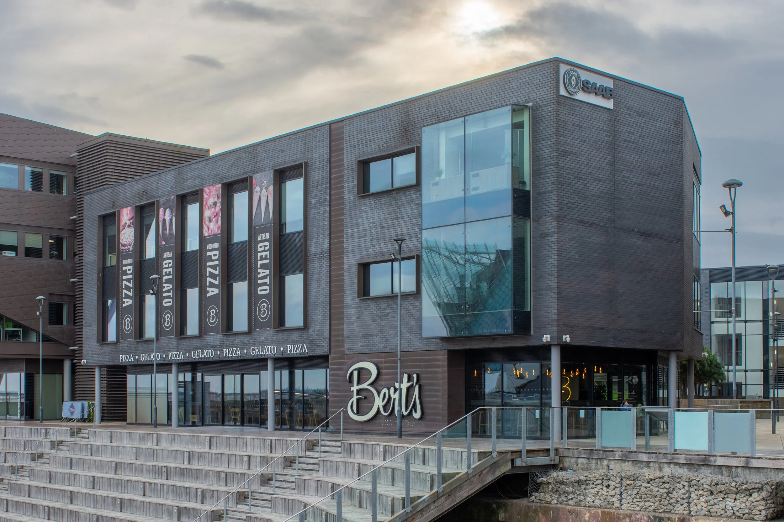 Modern commercial building with a brick exterior, large glass windows, and signage for a pizza, gelato, and dessert shop. Outdoor wooden boardwalk and stairs leading to the entrance, with a cloudy sky in the background.