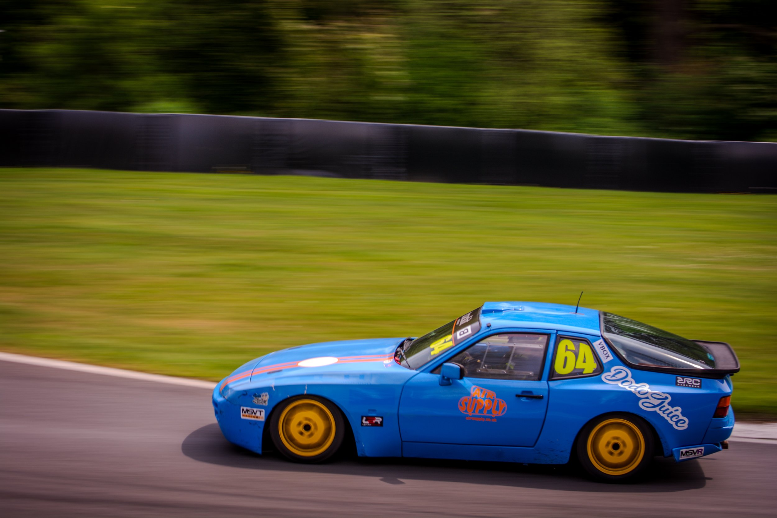 A blue race car with yellow wheels speeding along a track with a blurred background of greenery.