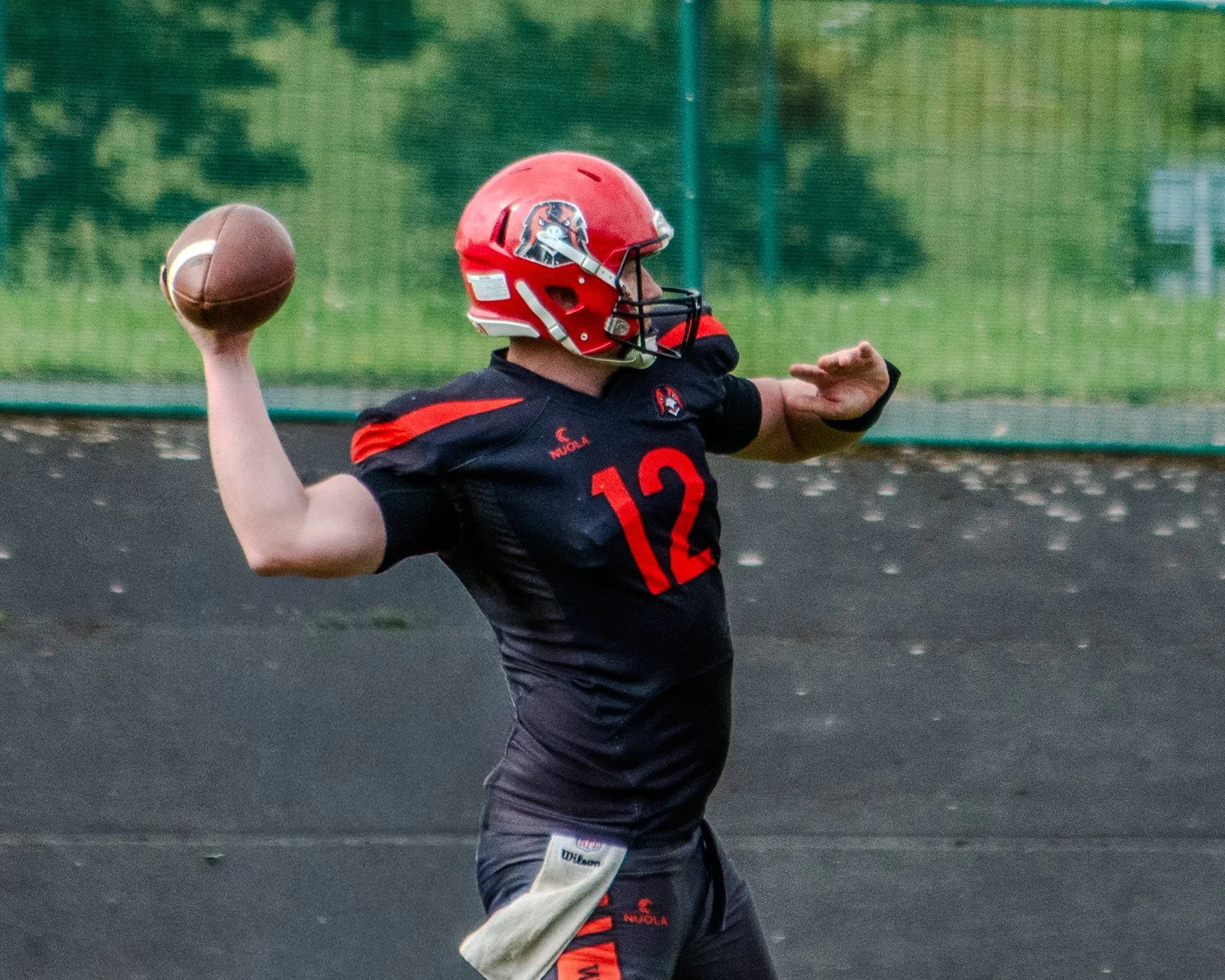 A football player is throwing a football on an outdoor field, wearing a red helmet and a black jersey with the number 12.