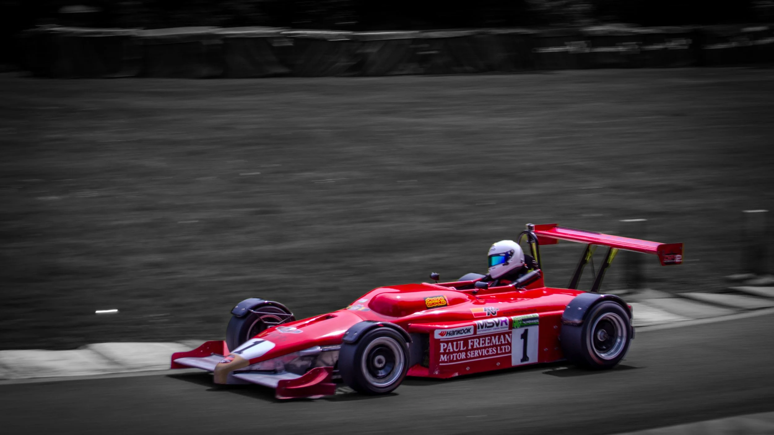 A red Formula race car with the driver wearing a white helmet racing on a track, with the background in black and white.