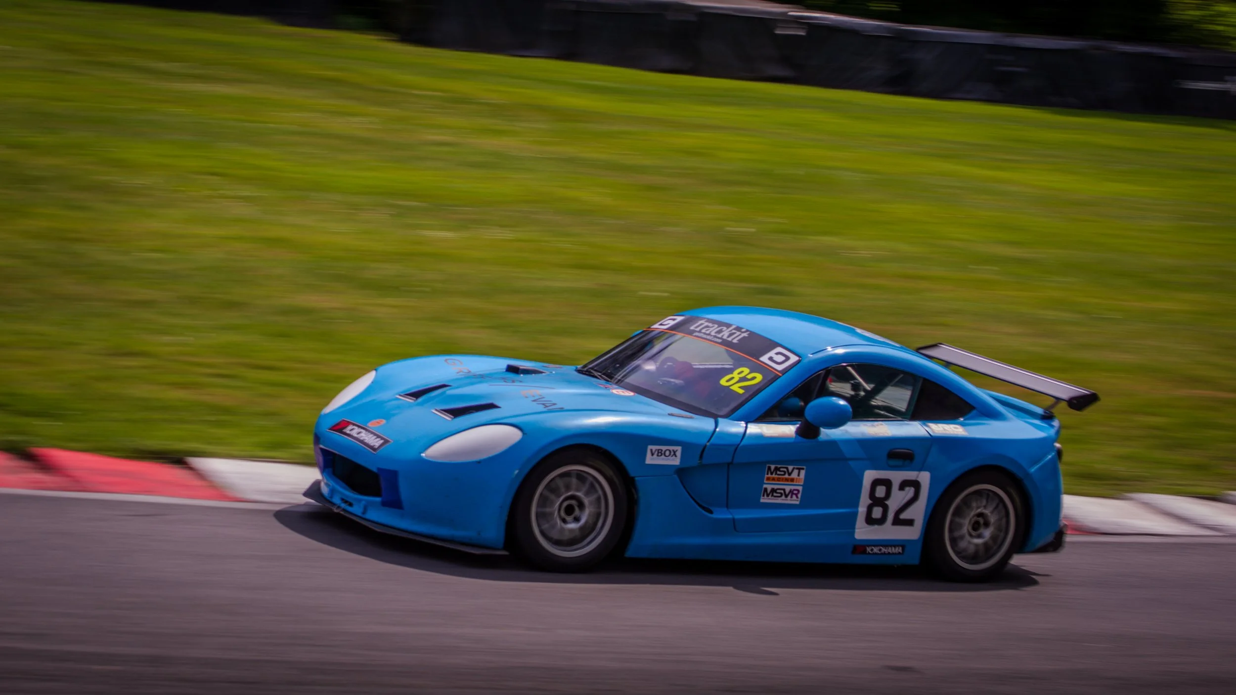 A blue race car with the number 82 on it speeding on a race track with a grassy background.