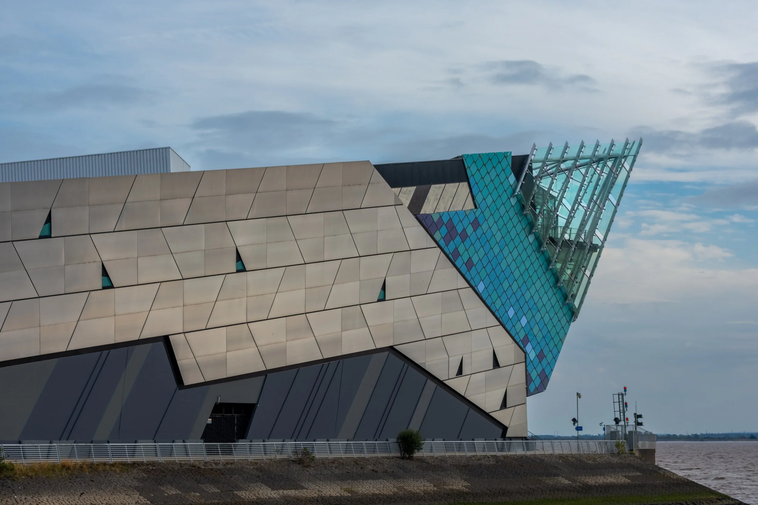 Modern building with angular design, featuring sections of beige panels, blue mosaic-like glass, and green-tinted glass structures against a partly cloudy sky.