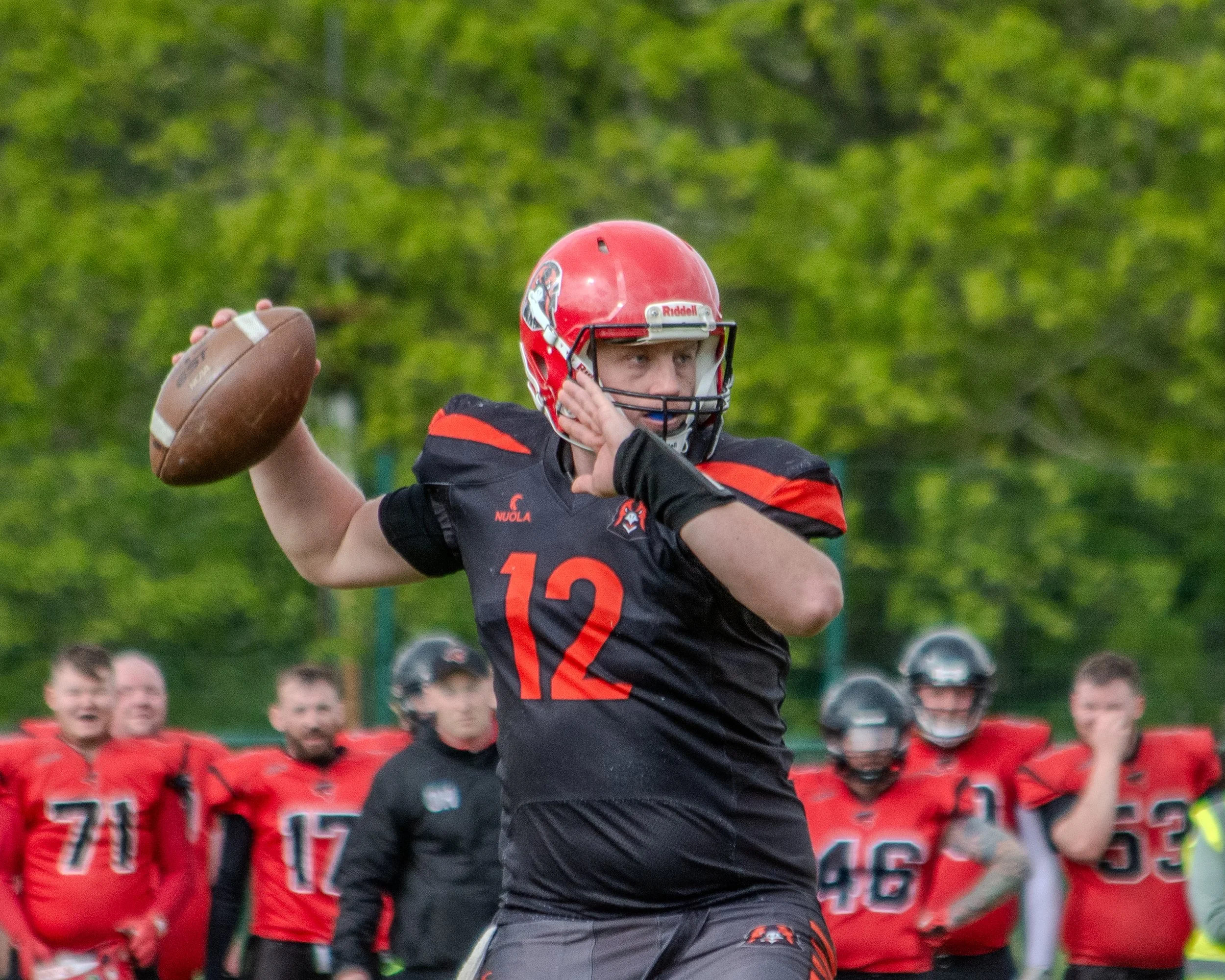 A football player in a black and red uniform, wearing a red helmet with a logo, is throwing a football during a game. In the background, there are team members in red uniforms and spectators.