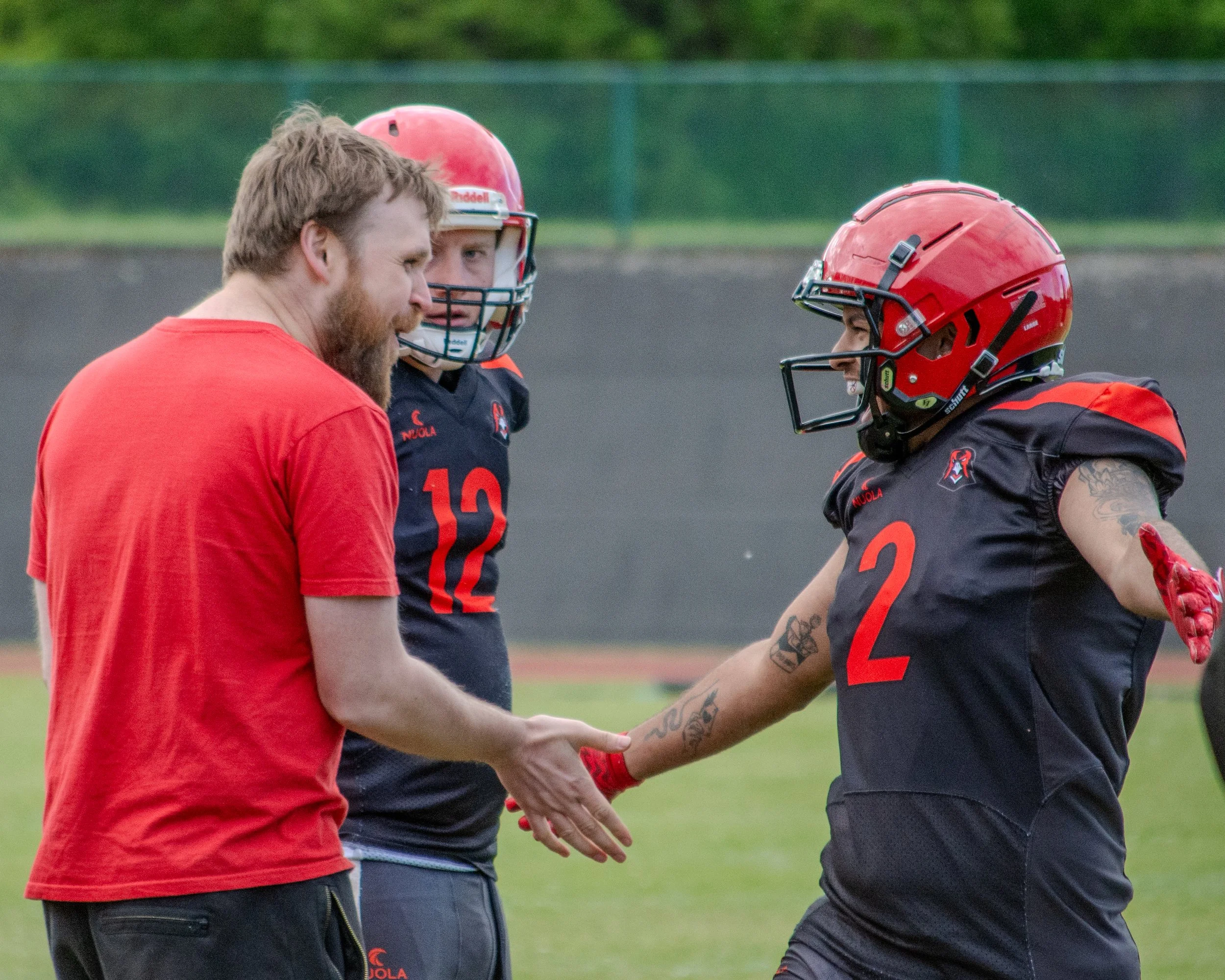 Two football players and a coach or trainer on a field. The players are wearing helmets and jerseys, and they appear to be greeting or congratulating each other while the coach looks on.