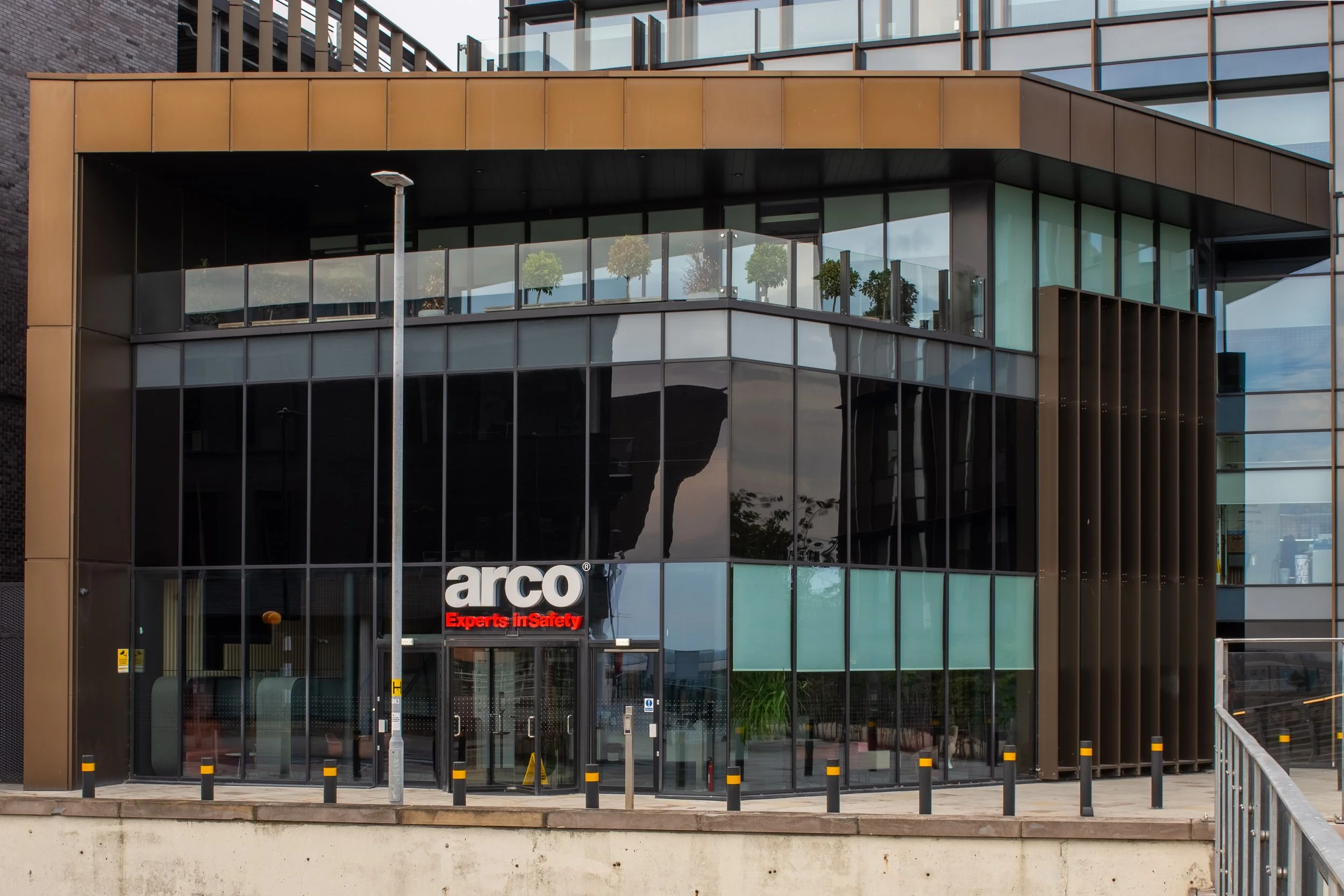 Modern commercial building with glass windows and a gold-colored facade, displaying the logo 'arco' and the slogan 'Experts in Safety'.