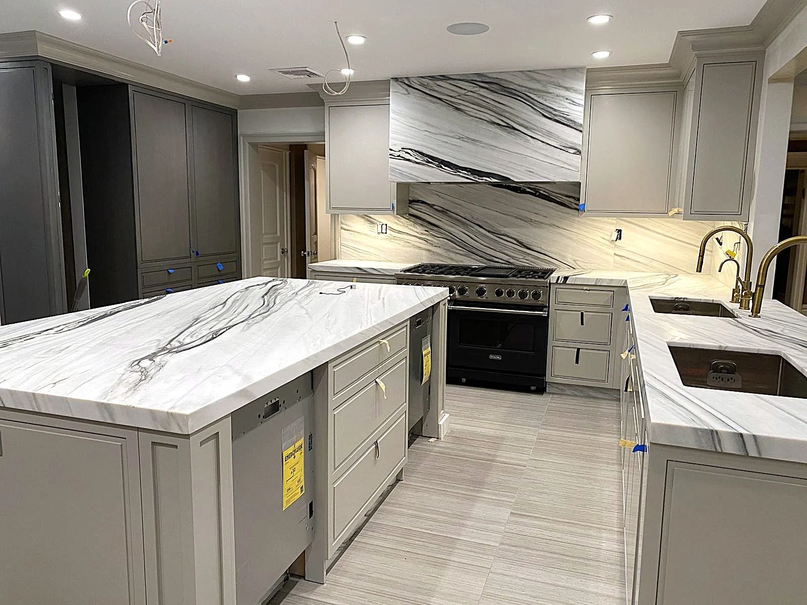 A kitchen under construction with gray cabinetry, white and gray marble countertops, a black stove, and a brass sink faucet.