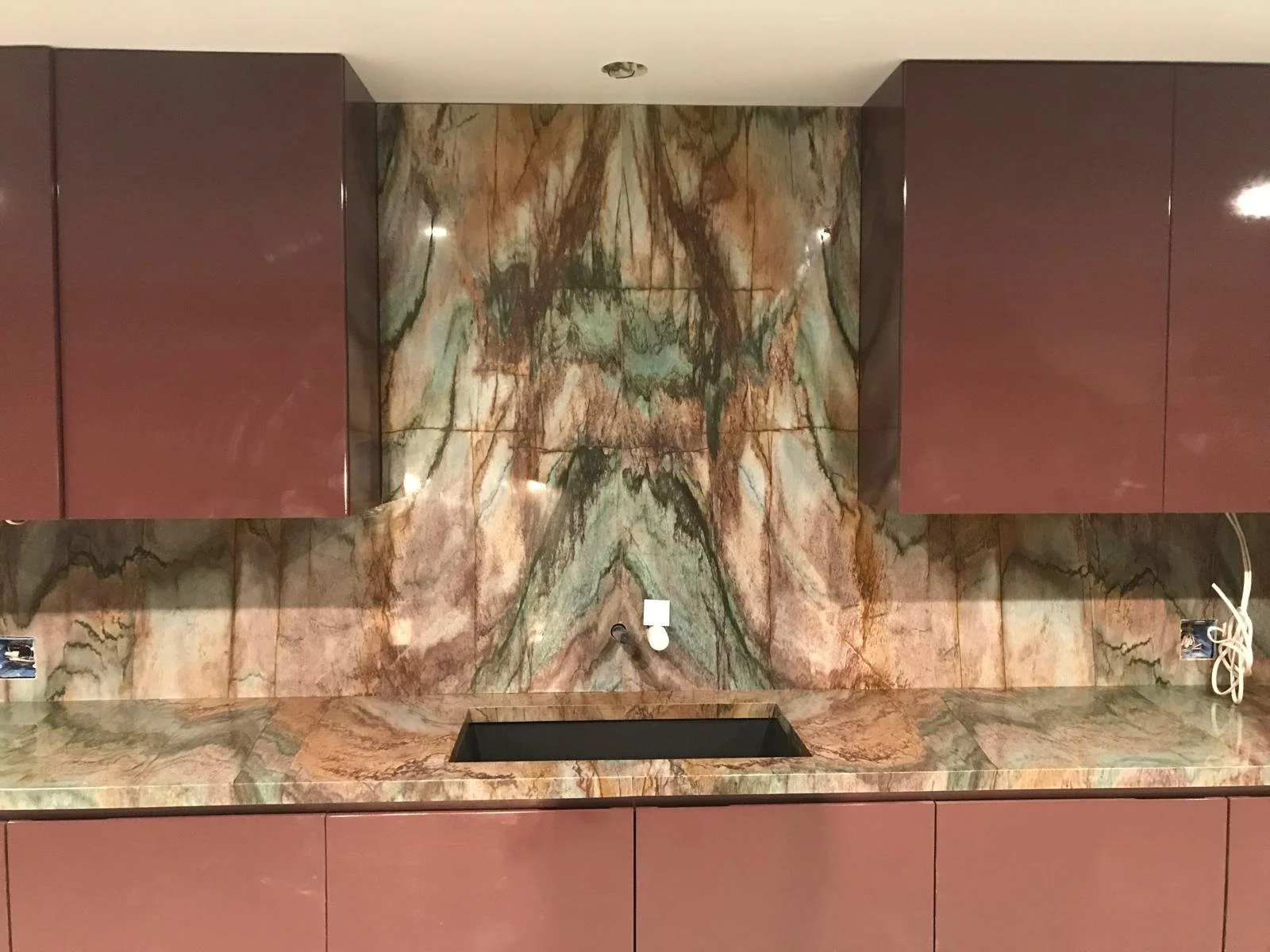A kitchen backsplash and countertop made of multi-colored marble with earthy tones and intricate veining patterns, topped with maroon cabinets.