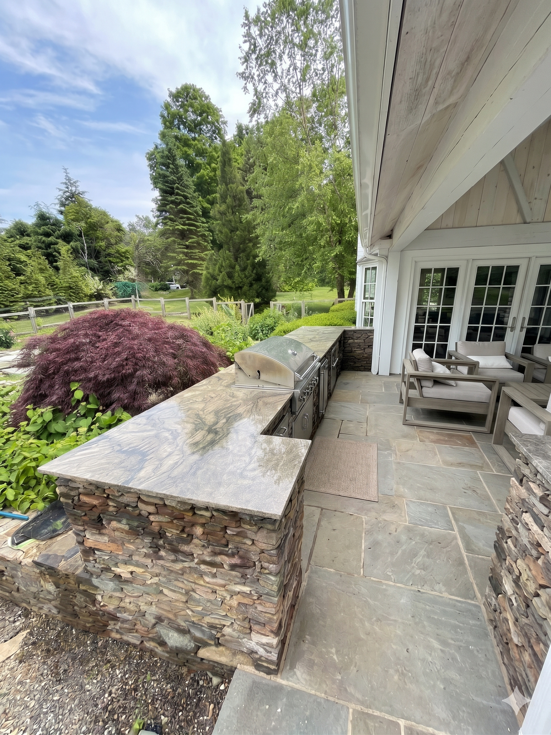 The image shows a backyard patio with a built-in outdoor kitchen area featuring a gas grill and stone counter. There are outdoor chairs and a table. The patio is paved with large stone tiles and is surrounded by lush greenery and tall trees, with a b