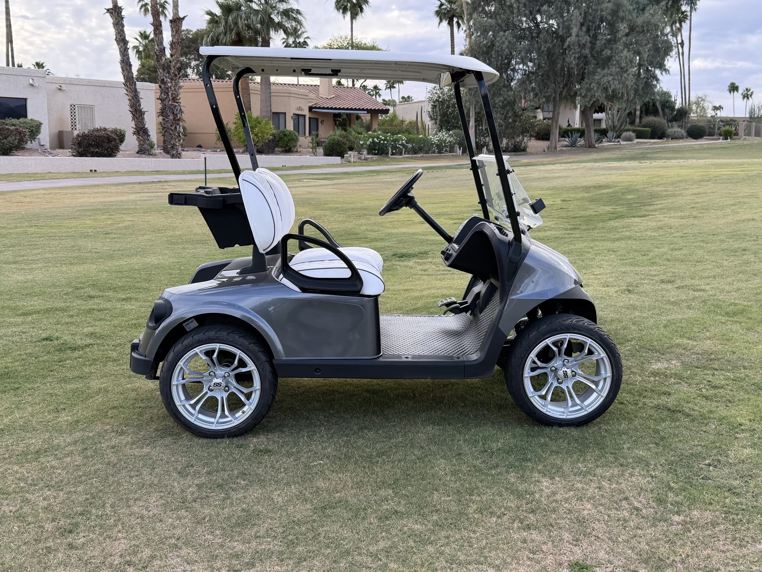 A golf cart with white seats and a black roof is parked on a well-maintained grassy area at a golf course, with houses and palm trees in the background. Rio Verde Golf Cart, Fountain Hills, Scottsdale Golf Cart, Scottsdale golf cart repair