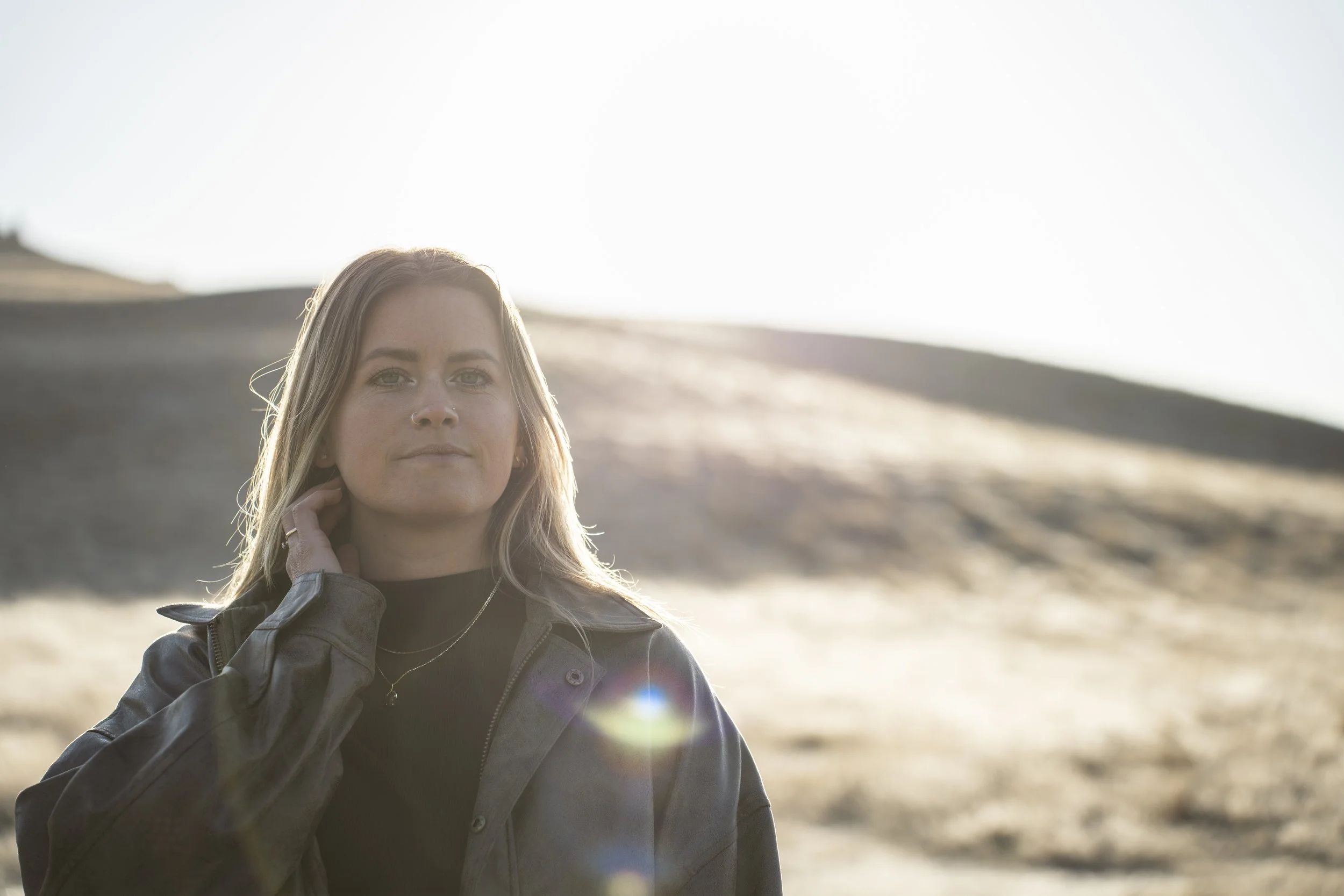 Portrait of a young woman with blonde hair and freckles, wearing a black top and a gray jacket, standing outdoors during sunset with hills in the background.