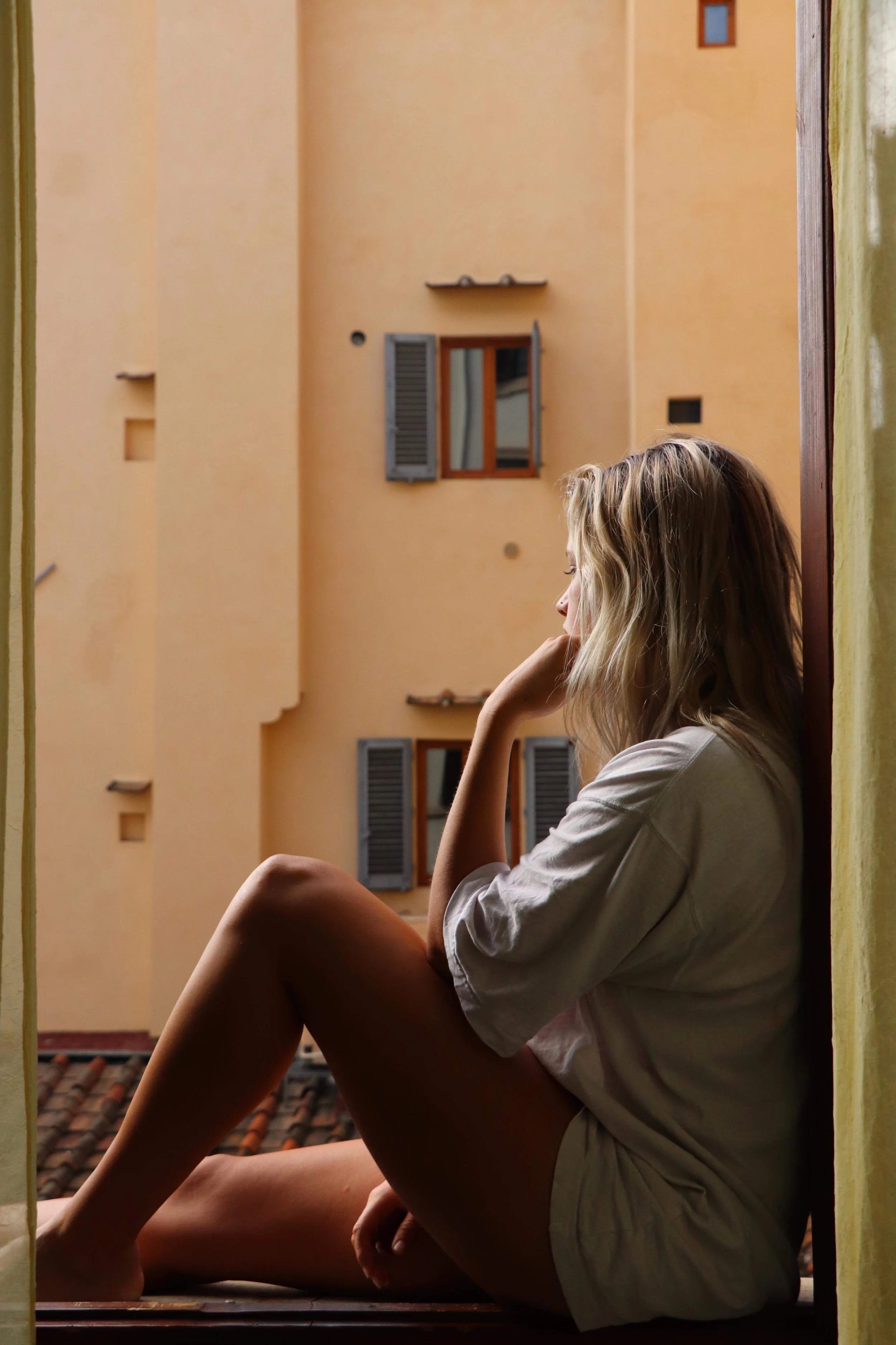A woman with long blonde hair sitting on a balcony, looking thoughtful, with their eyes closed, in front of an orange building with windows and shutters.
