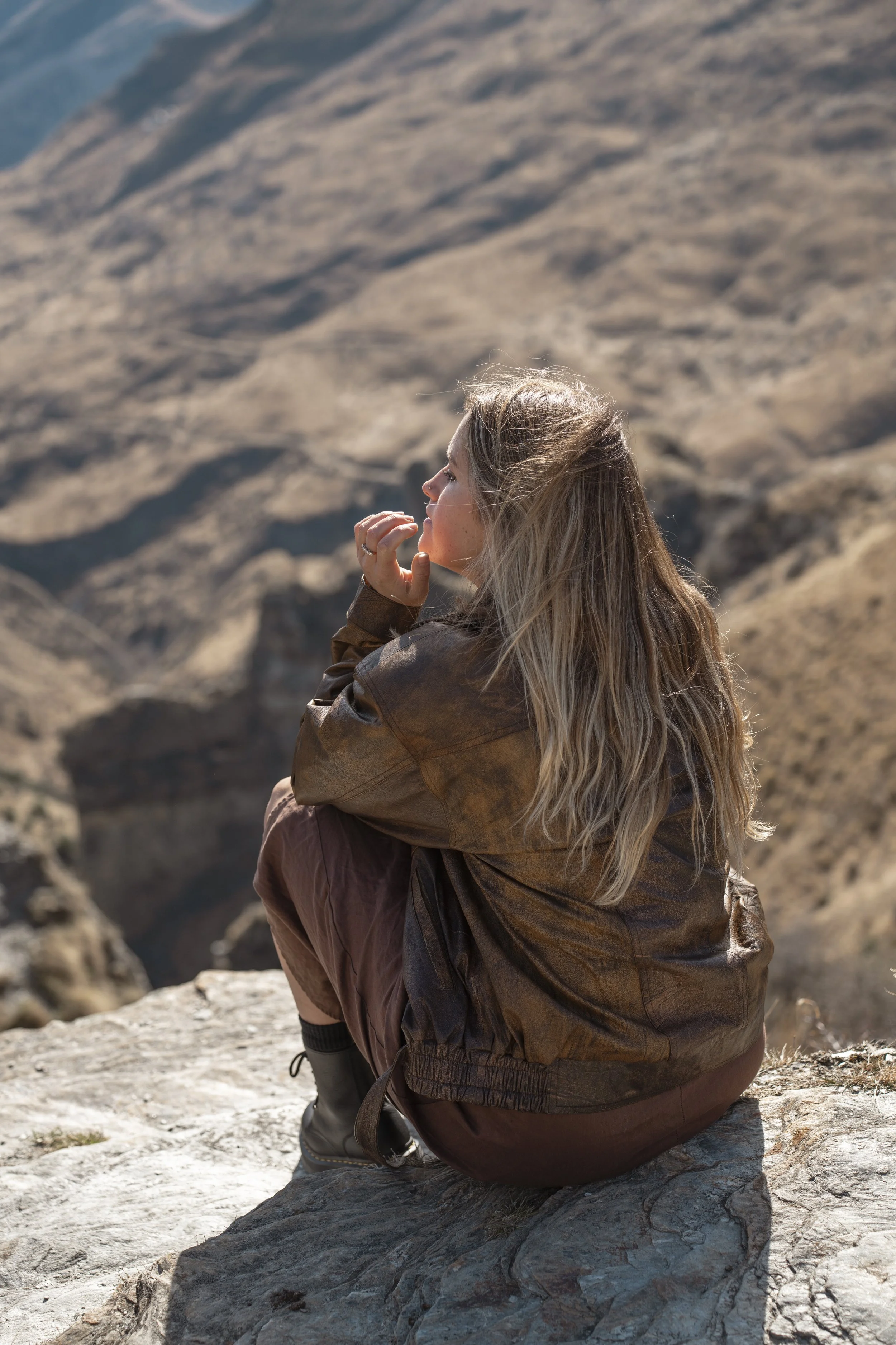 A woman with long blonde hair sitting on a rock in a desert landscape, wearing a brown jacket and maroon pants, with mountainous terrain in the background.
