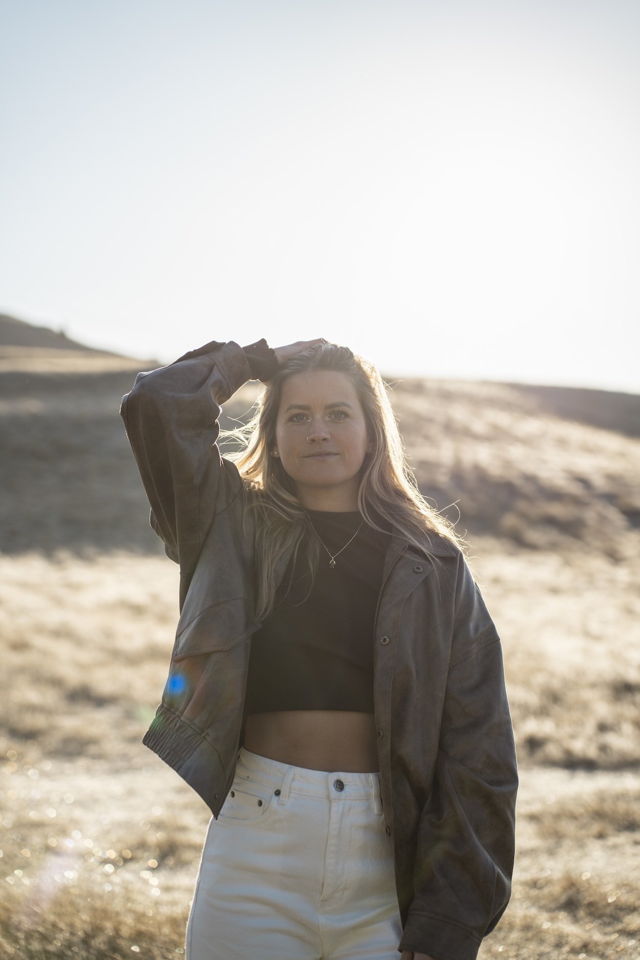 Kelsey standing outdoors on a sunny day, wearing a black crop top, a black leather jacket, and white pants, with her hand resting on her head and a natural landscape in the background.