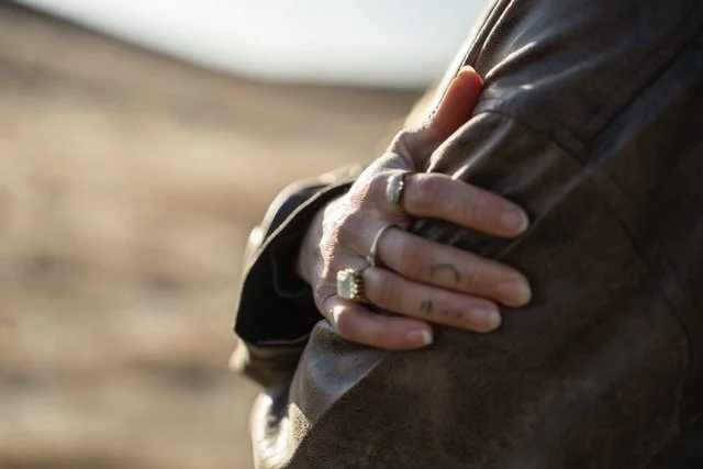 A close-up of a person's hand with multiple rings resting on their arm, wearing a dark leather jacket, with a blurred outdoor background.