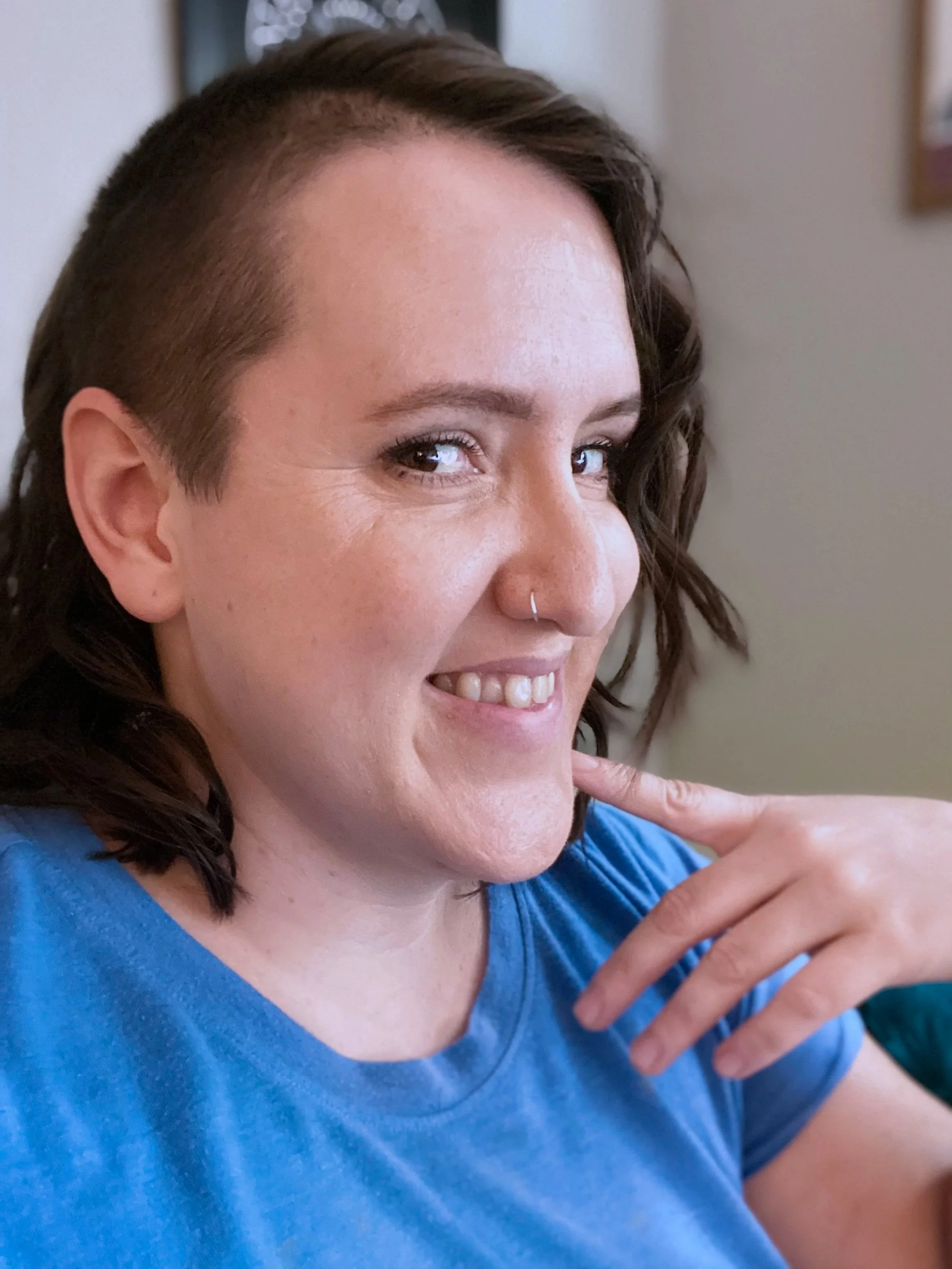Close-up of Crystal with brown curly hair, a nose ring, and a blue shirt, smiling and pointing to herself.