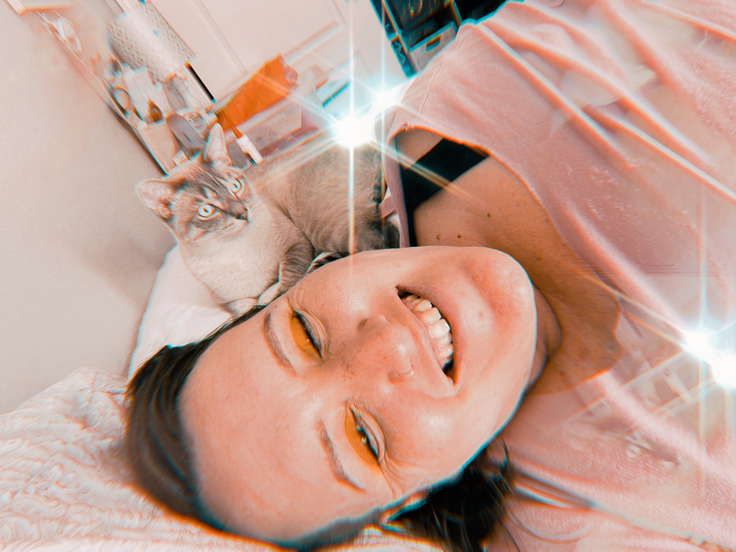 Crystal lying on a bed, smiling with her eyes closed, and a tabby cat sleeping on her chest. The room has a cabinet and various items on shelves, with a bright and relaxed atmosphere.