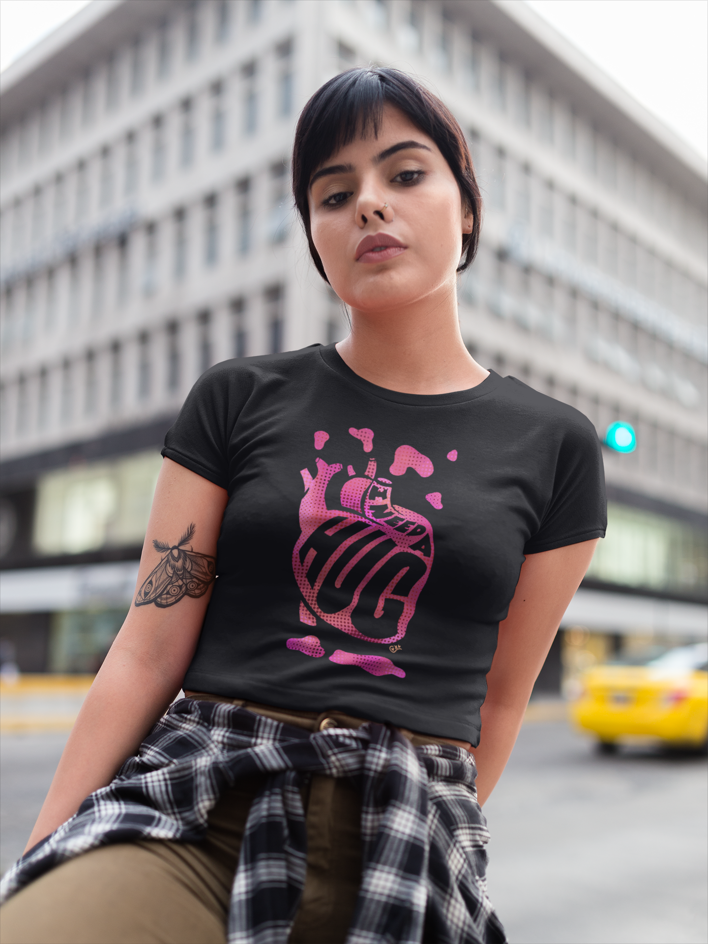 A young woman with short dark hair and a nose piercing poses in an urban setting. She wears a black t-shirt with pink graphic text and has a plaid shirt tied around her waist. A city building and yellow taxi are visible in the background.