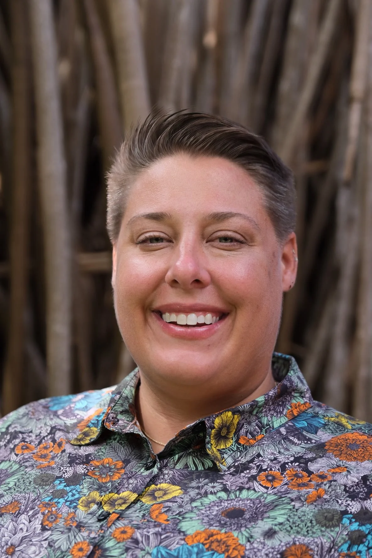 A smiling woman with short, styled hair, wearing a colorful floral-patterned shirt, standing in front of a rustic wooden background.
